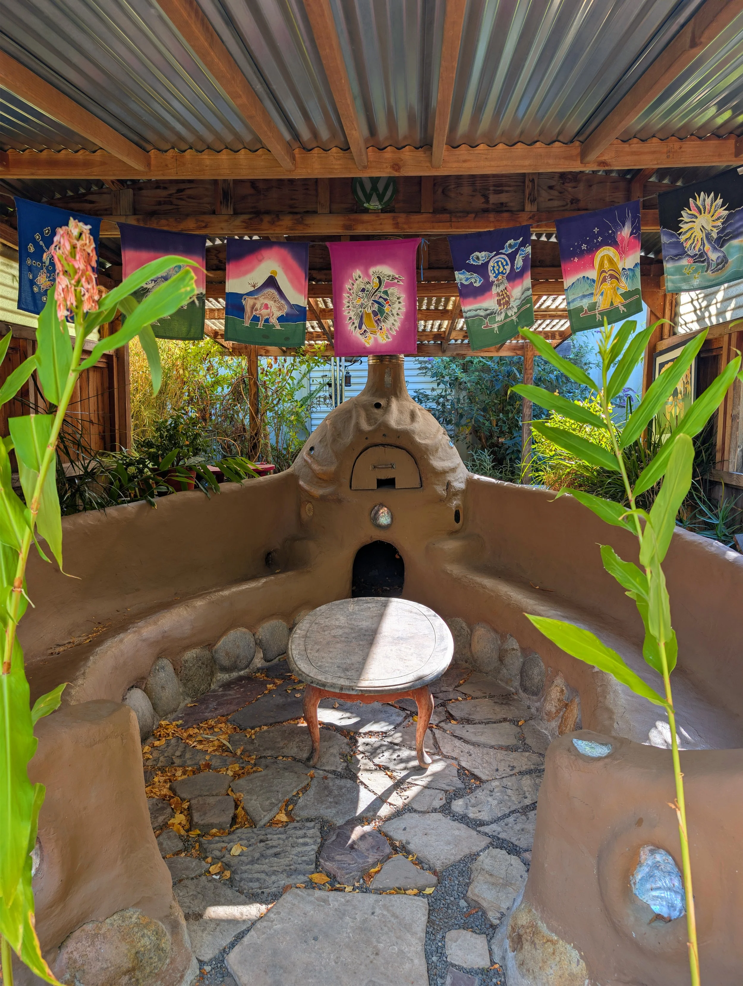 Outdoor area with adobe-style seating, a clay oven shaped like a turtle, surrounded by green plants, and decorated with colorful flags hanging from a wooden roof.