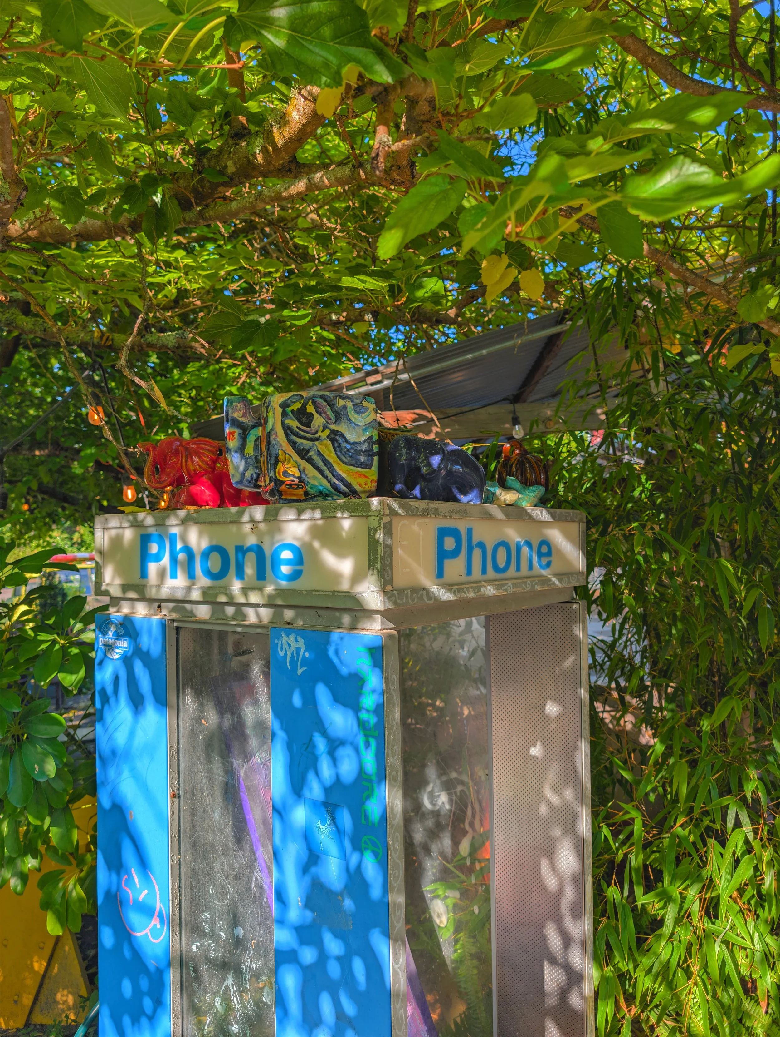 Outdoor scene of a payphone booth surrounded by lush green tree foliage. Tucked on top of the booth are holiday-themed ceramic figures and colorful art pieces, with sun and shadows creating a vibrant, lively atmosphere.