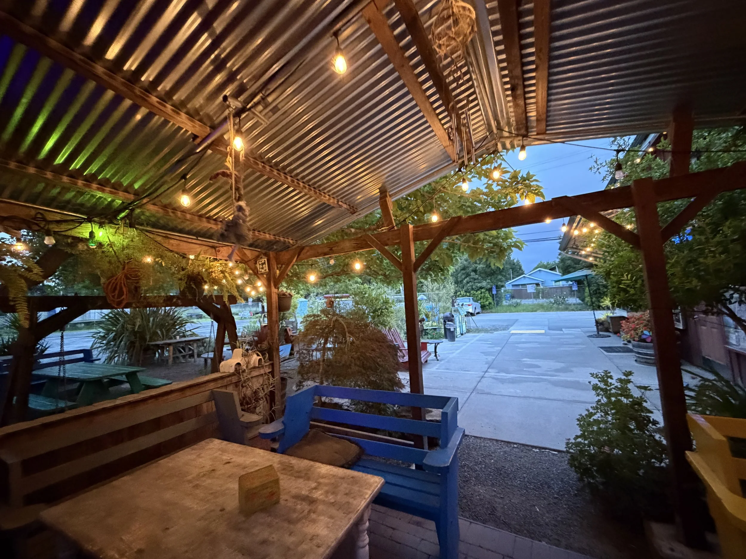 An outdoor patio with wooden tables and colorful chairs, string lights hanging from a metal roof, trees, a street with parked cars, and a house in the background, captured during evening twilight.