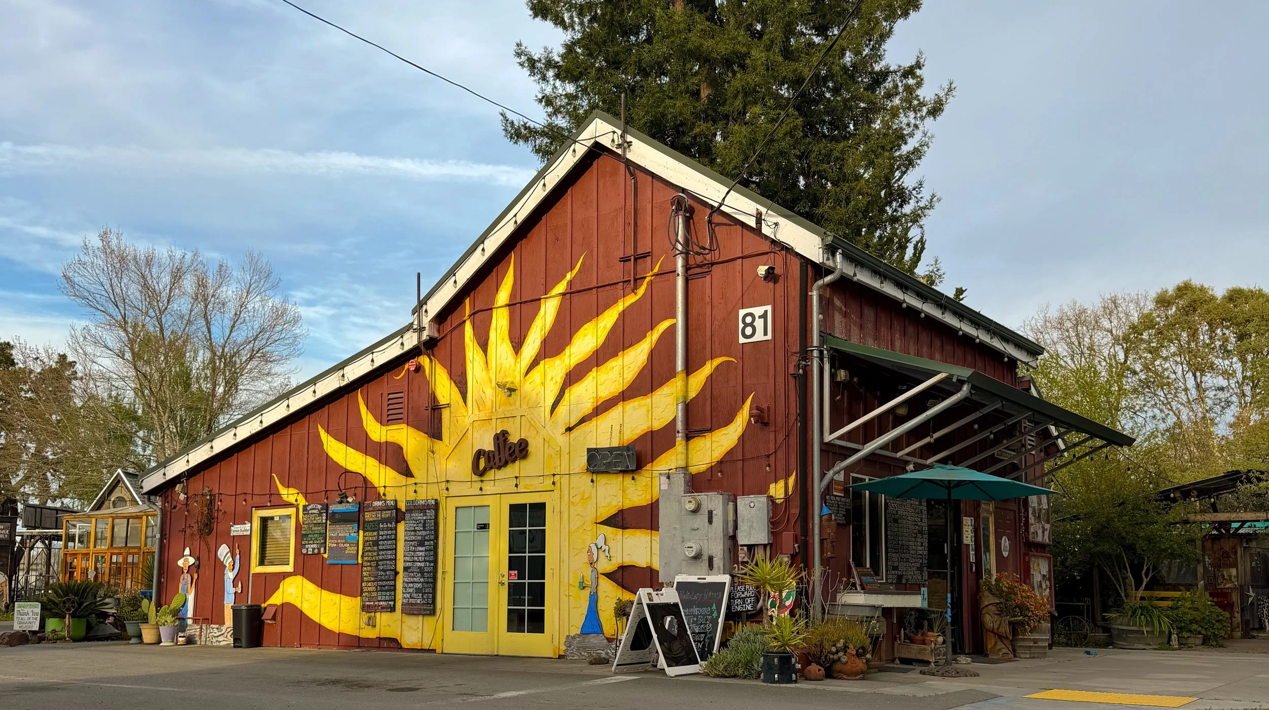 A red building with a yellow sunburst mural painted on it, with the word 'coffee' and menu boards on the front. There are potted plants and outdoor seating in front, with trees and a cloudy sky in the background.