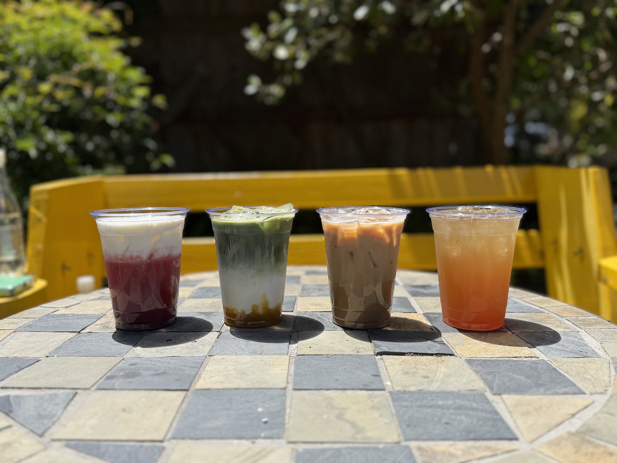 Summer seasonal cold beverages on a mosaic table outdoors with yellow benches in the background and trees with green leaves.