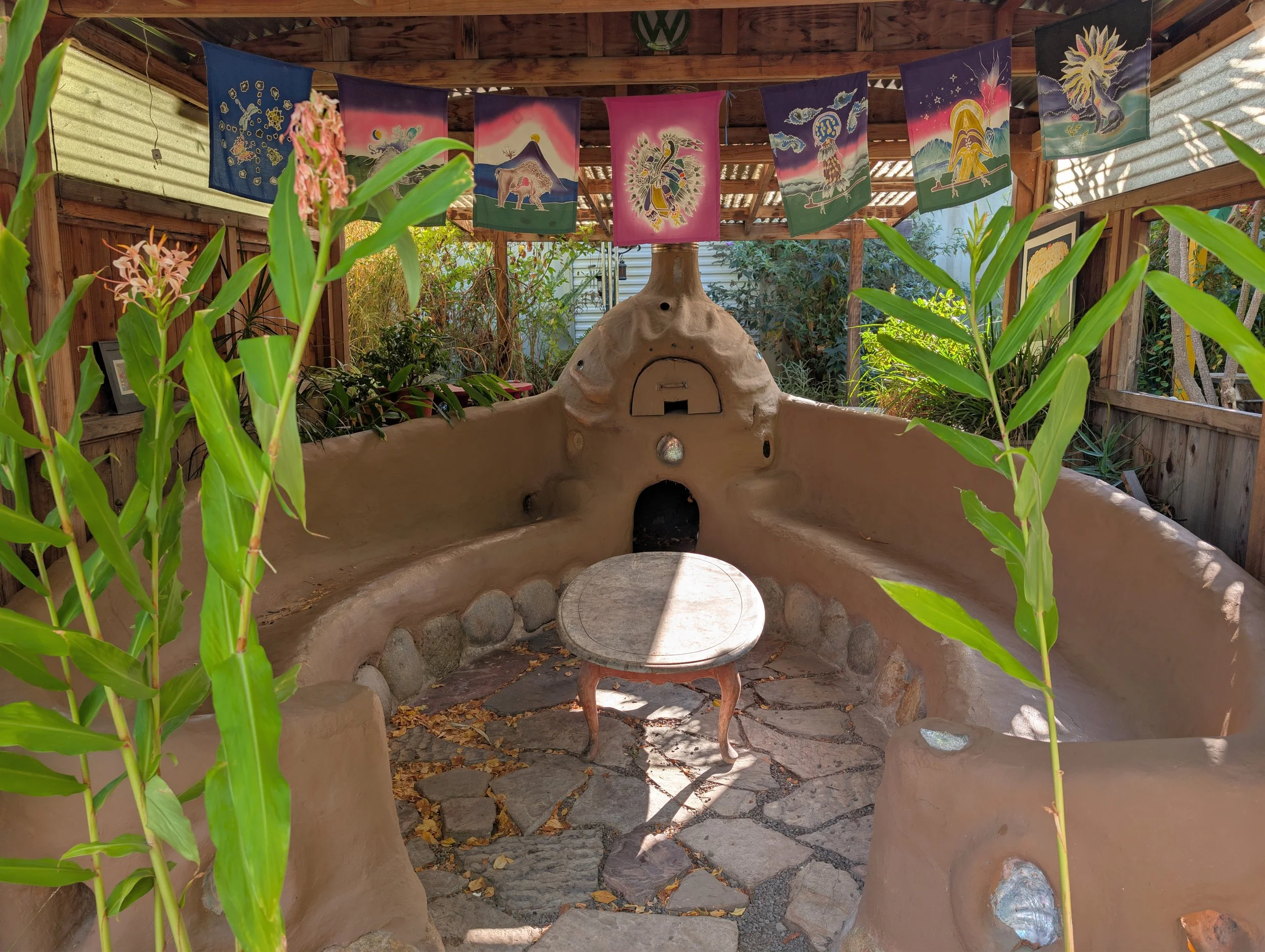 Outdoor patio area with a clay oven, surrounded by green plants and colorful hanging banners with artistic designs.