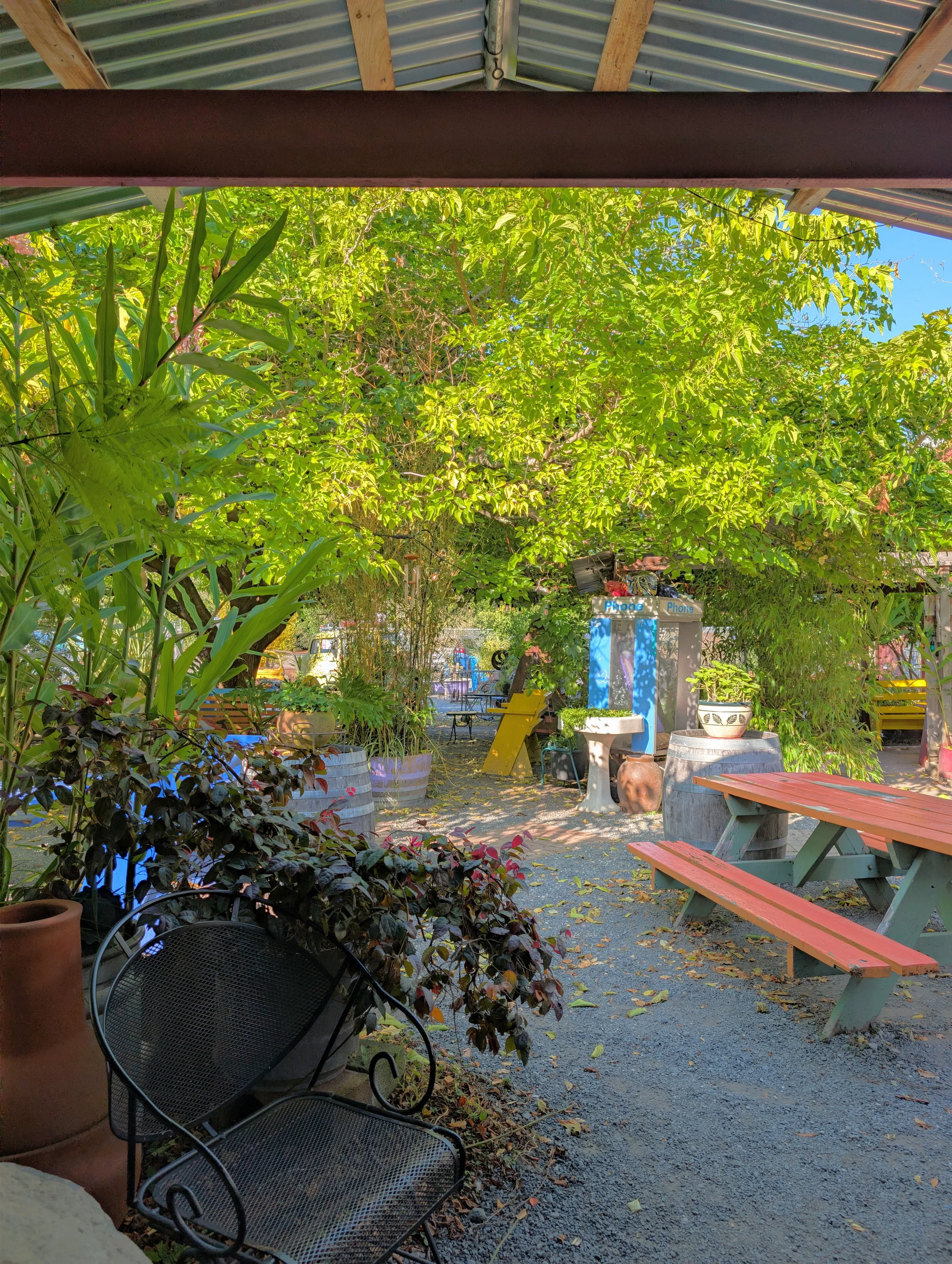 An outdoor cafe or garden seating area with black wrought iron chairs and a red and green picnic table. The area features lush green trees and plants, a blue phone booth, and a gravel ground with fallen leaves. The space is covered by a metal roof su