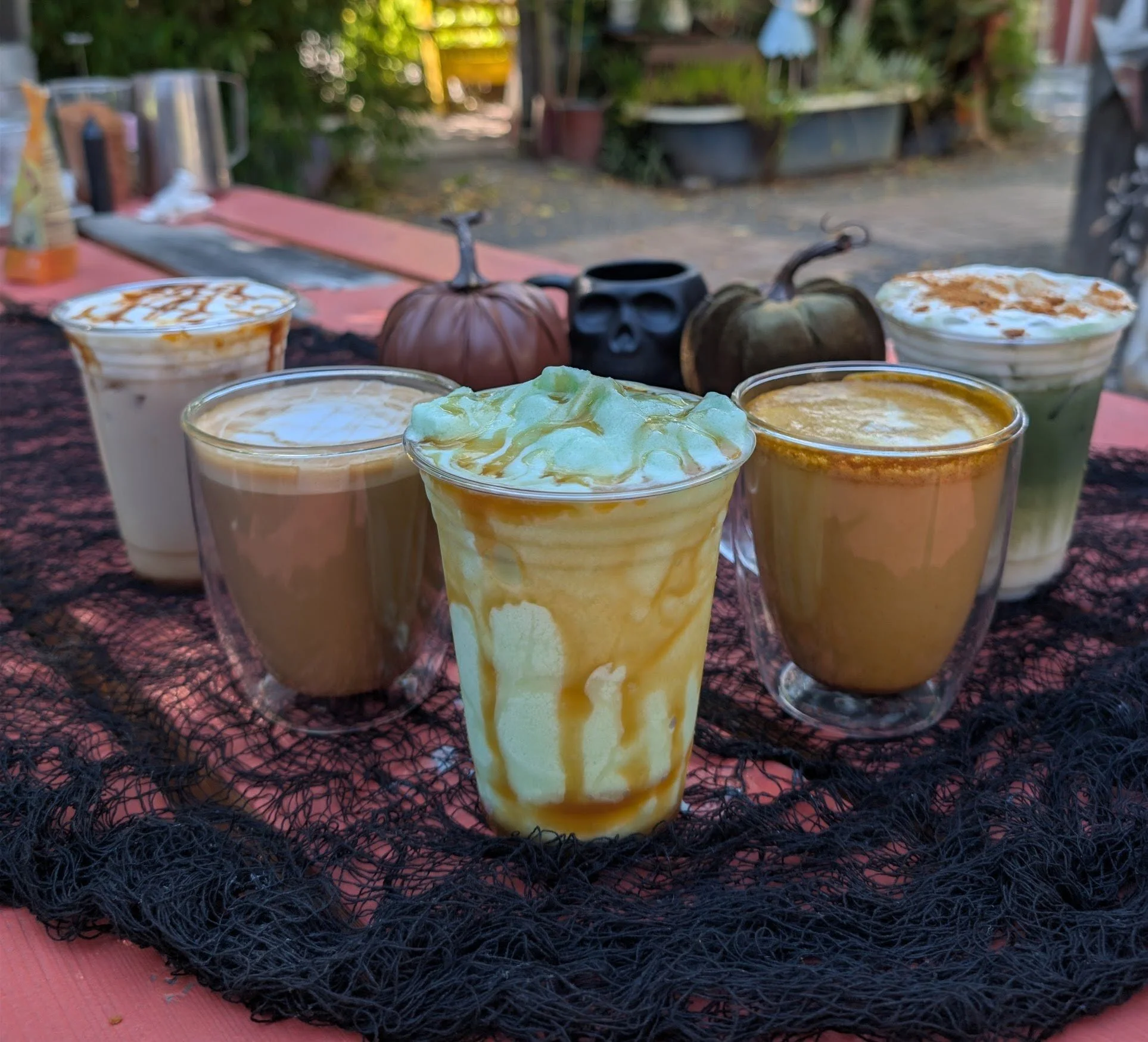 Five drinks with whipped cream and caramel sauce on top, placed on a black netted surface, with Halloween-themed pumpkins and skull cups in the background.