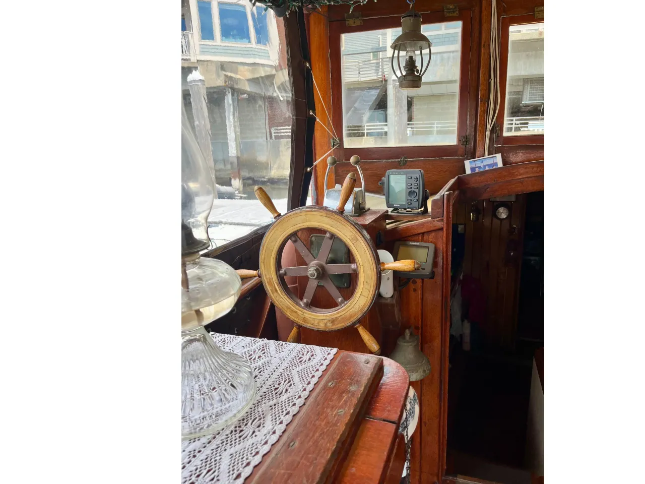 Close-up view of boat's wooden steering wheel with antique navigation instruments and digital gauge, inside a boat cabin with large windows and a hanging lantern.