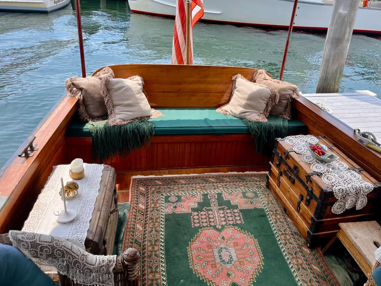 A cozy boat deck with a wooden bench, beige pillows, and green throw blankets. There is a small side table with a candle and a decorative item, an ornate rug on the floor, and a wooden chest with a doily and a plate of strawberries. Water with boats in the background.