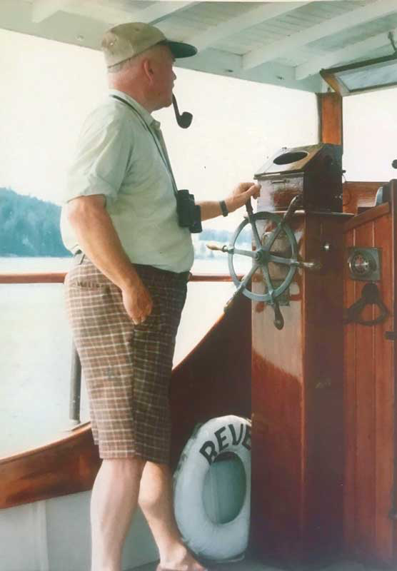 A man standing on a boat bridge with a pipe in his mouth, wearing a hat, sunglasses, a watch, and plaid shorts, looking at a boat's steering wheel and controls, with a life preserver labeled 'REVE' on the deck.