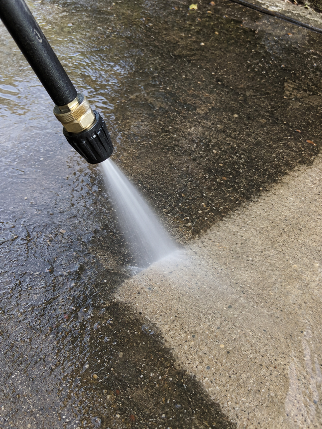 A close-up of a hose spray nozzle washing a concrete surface outdoors with water.