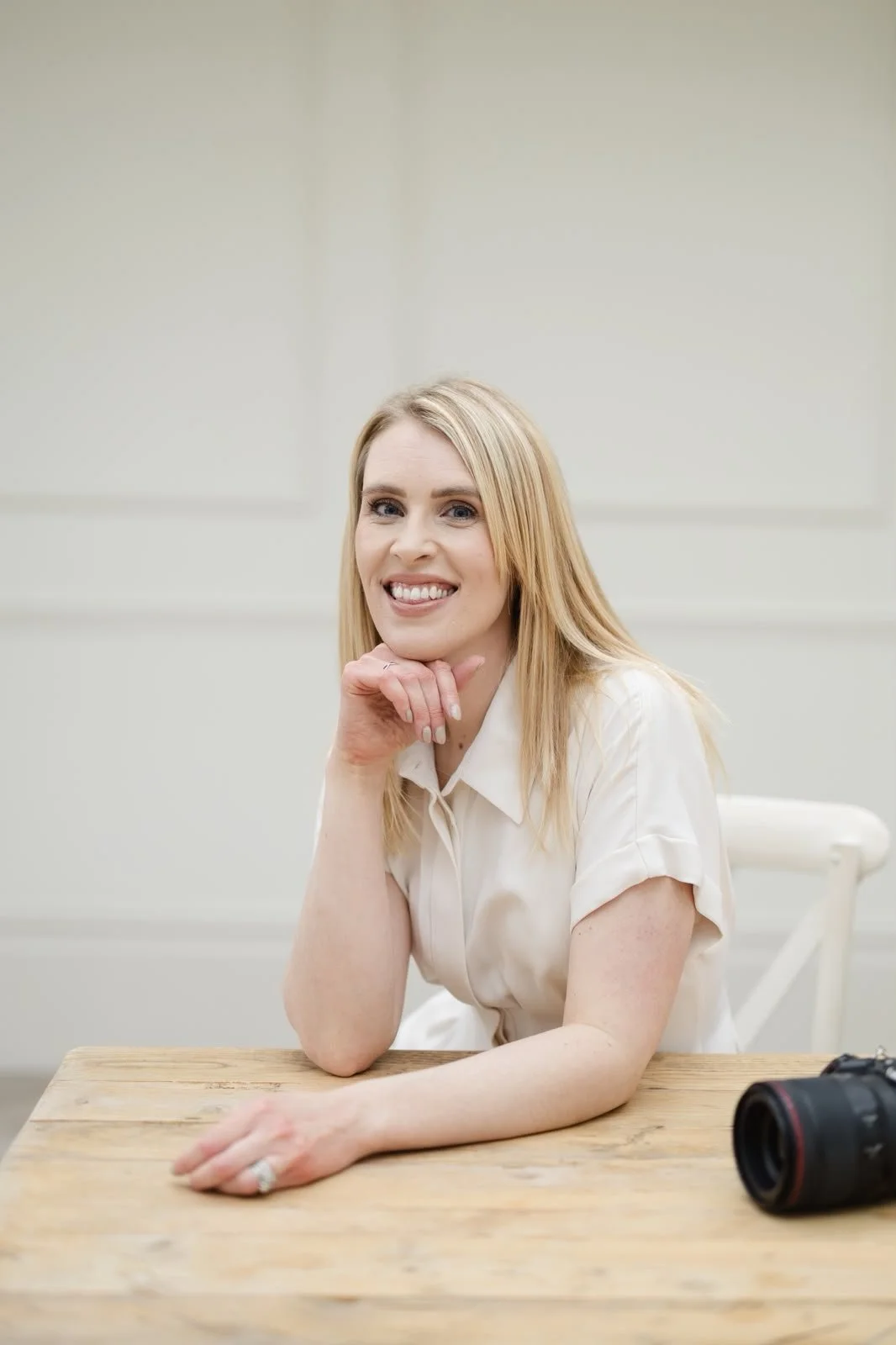 A woman with blonde hair smiling and resting her chin on her hand, sitting at a wooden table with a camera on it.