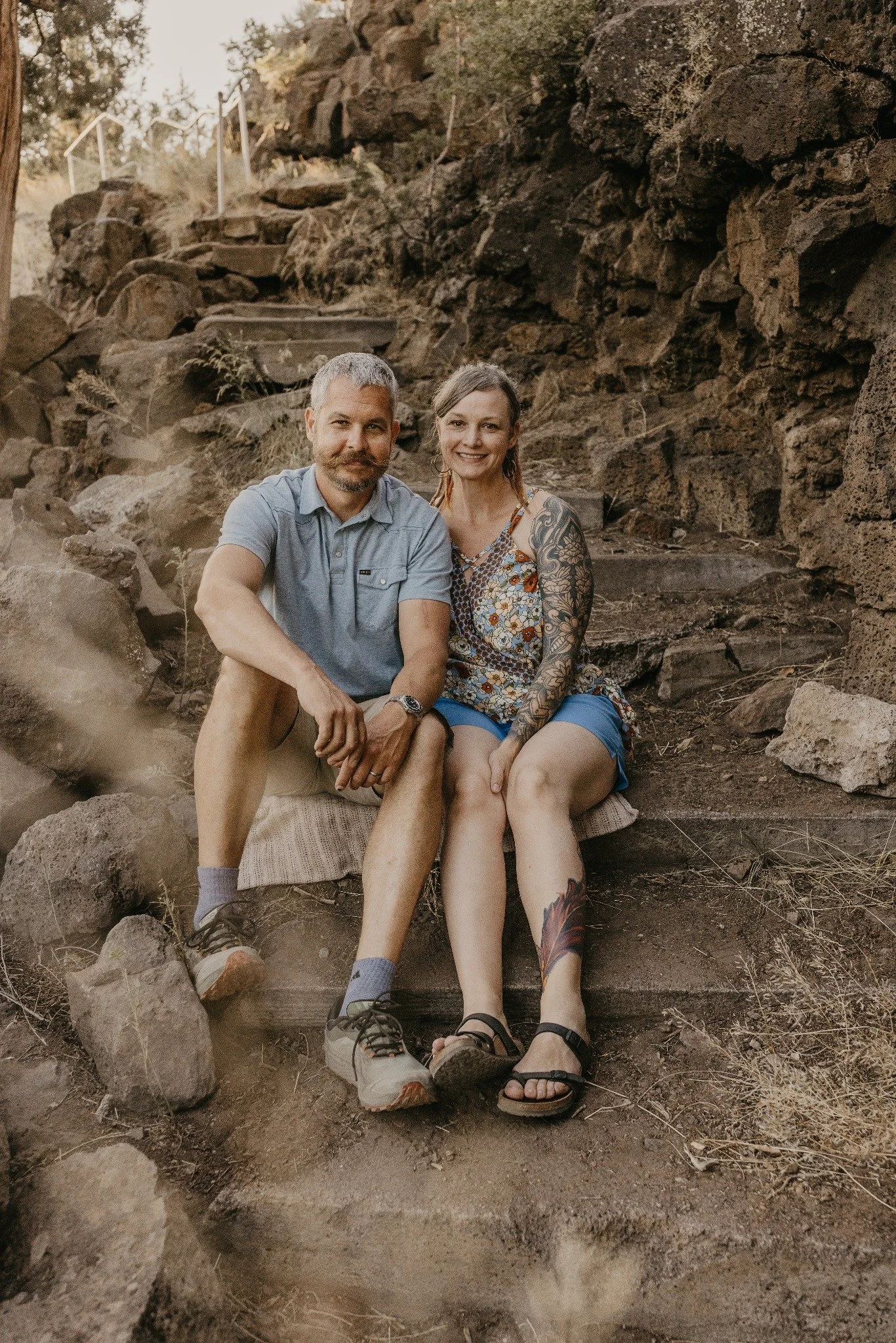 A man and woman sitting on a wooden bench outdoors next to a rocky hillside. The man has short gray hair, a beard, and is wearing a blue shirt and shorts. The woman has long hair, tattoos on her arms, and is wearing a floral top and shorts. They are smiling and relaxed, with the landscape behind them.
