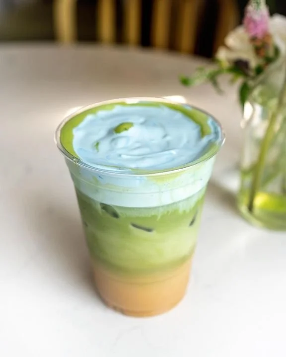 A layered beverage in a clear plastic cup with green, white, and brown layers, topped with a frothy blue-green foam, sitting on a white surface with a blurred floral arrangement in the background.
