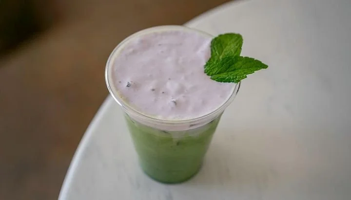 A tall glass of purple and green smoothie garnished with a mint leaf, on a white surface.