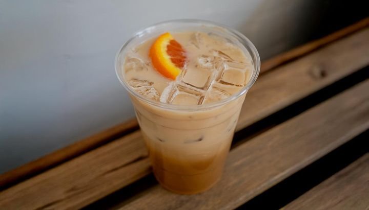 A clear plastic cup filled with iced coffee and ice cubes, garnished with a slice of orange, placed on a wooden surface.