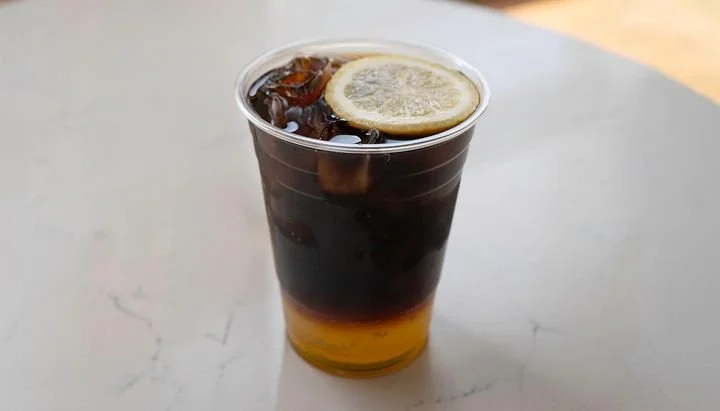 A plastic cup of iced black soda with ice cubes and a lemon slice on top.