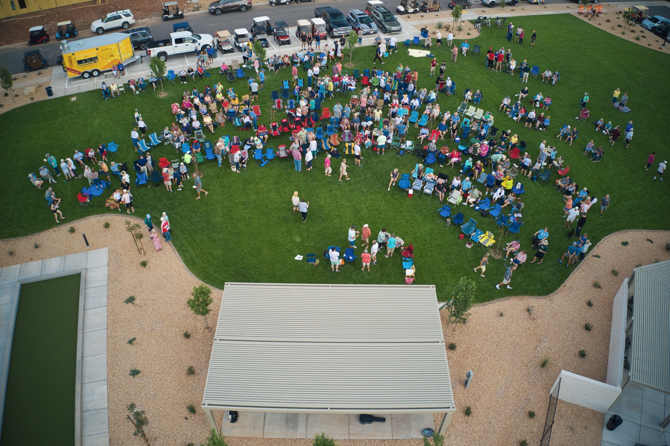 An aerial view of a large group of people gathered in a grassy area with many chairs, some filled, and some empty, during daytime. People are standing, sitting, and walking around. There is a row of parked cars along a paved parking lot border in the background. The area is part of a public event or gathering.