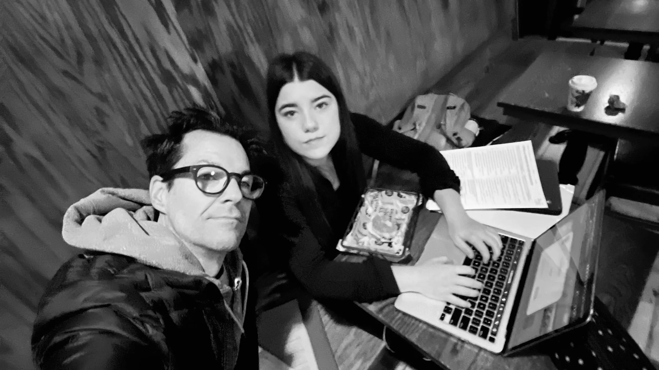 A black-and-white photo of a young man wearing glasses and a hoodie, and a young woman with long dark hair, sitting at a table with a laptop, a newspaper, and a drink. They are indoors, possibly in a cafe or restaurant.