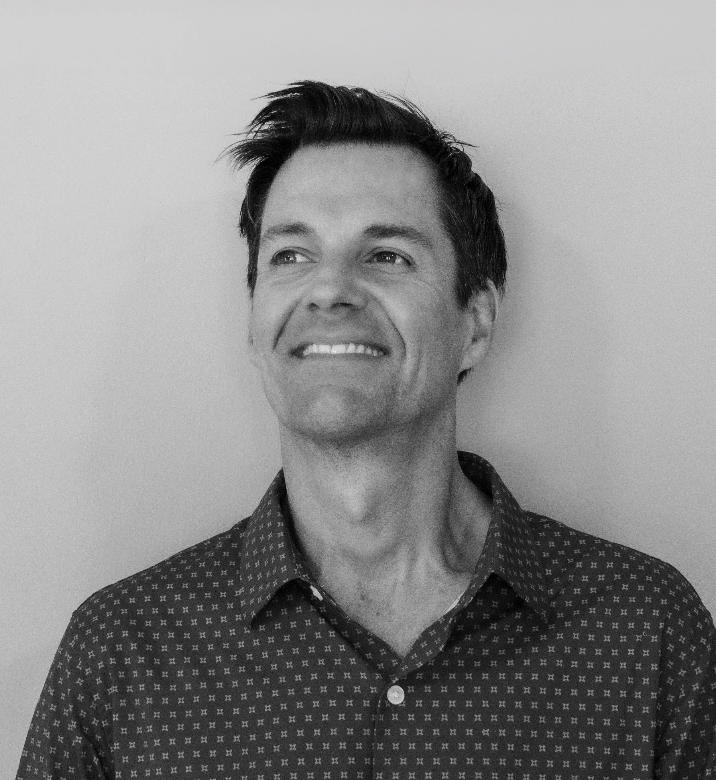 Black and white photo of a man smiling, showing only the top part of his face and his upper body, wearing a patterned shirt, with a plain background.