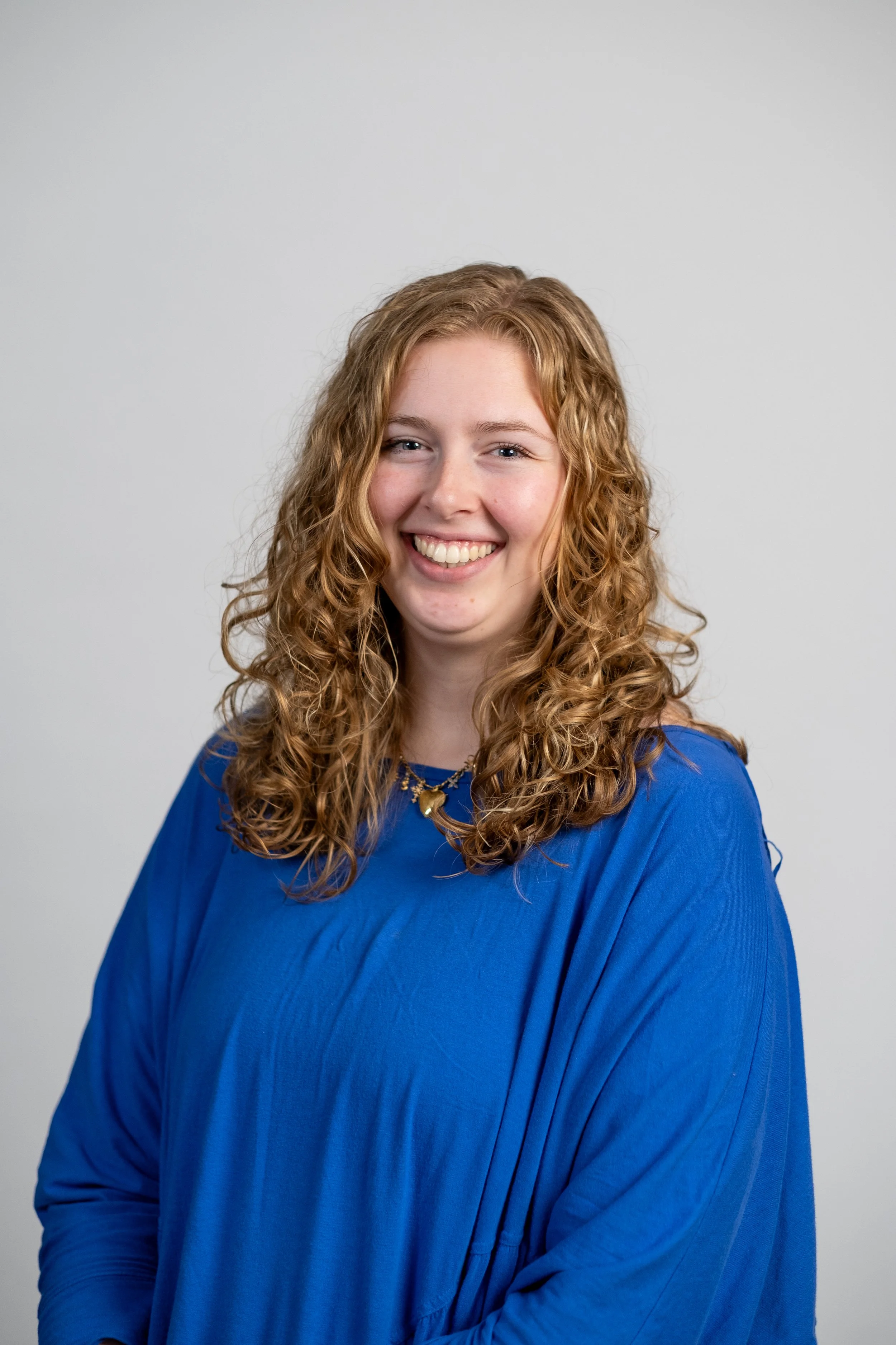 A young woman with curly red hair smiling, wearing a blue top and a gold necklace, standing against a plain white background.
