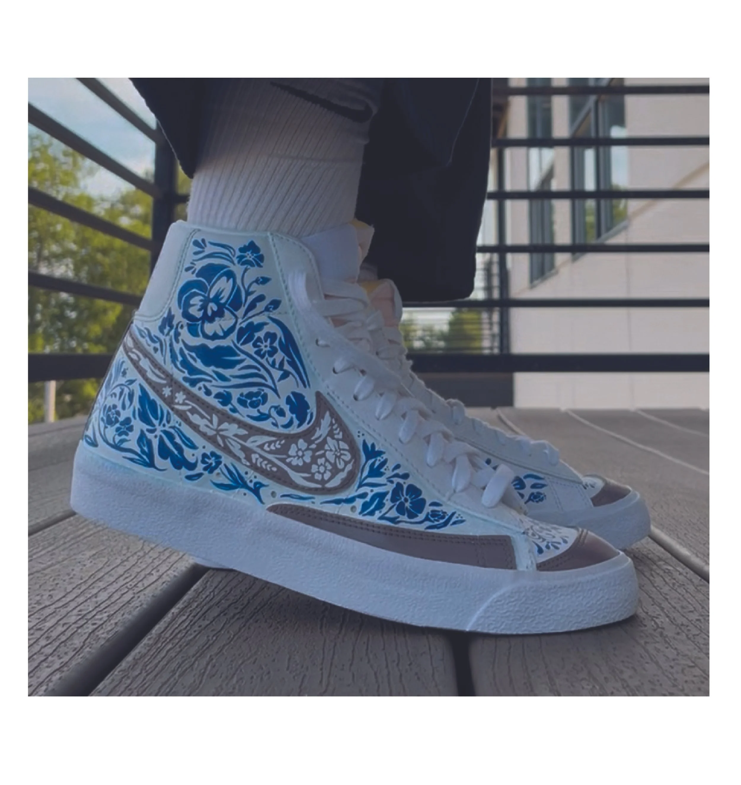 Close-up of white sneakers with blue floral designs on a wooden porch, with black railings and greenery in the background.