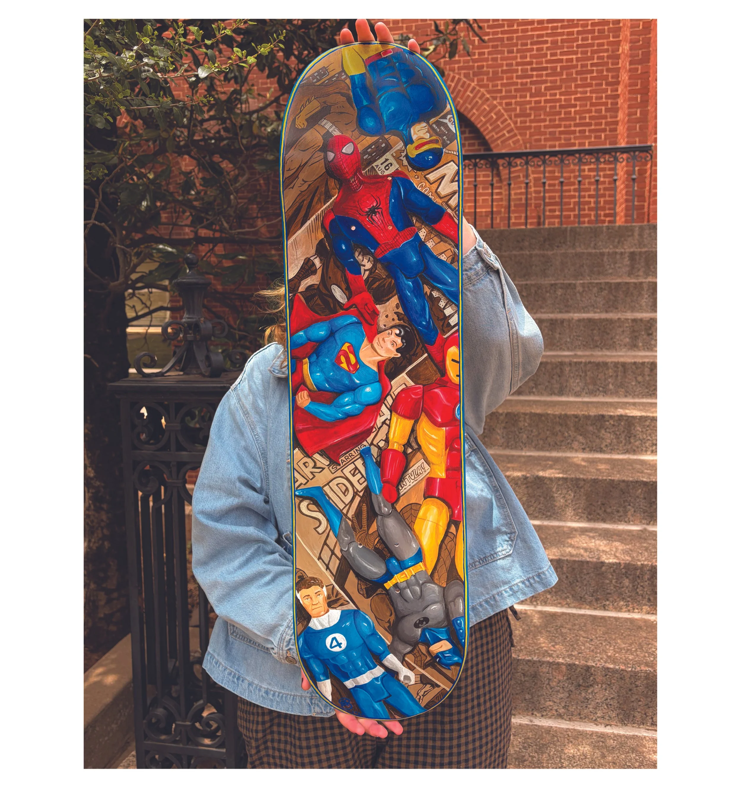 Person holding a skateboard with superhero illustrations of Spider-Man, Superman, and other comic characters in an outdoor urban setting.
