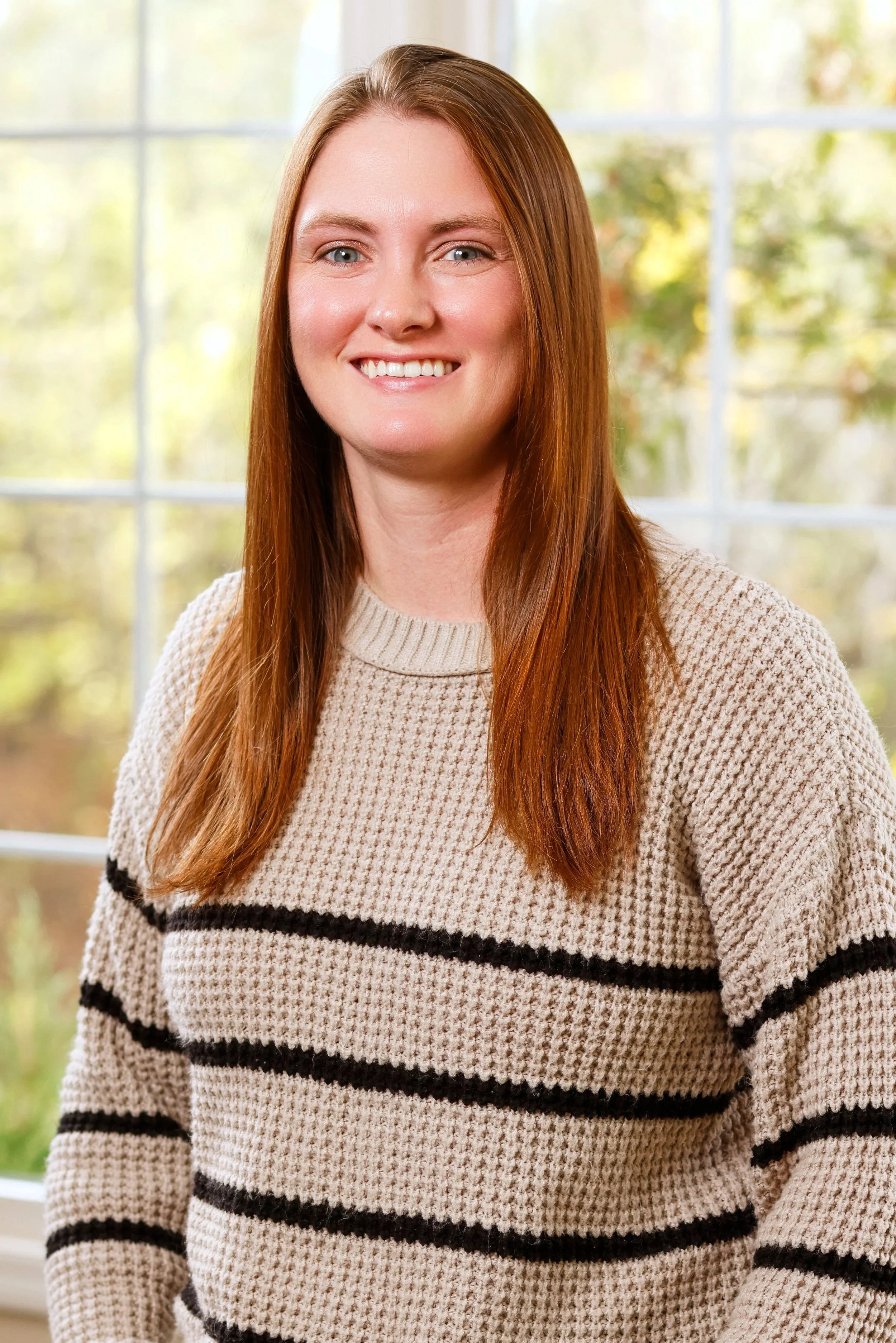 A woman with long red hair, blue eyes, and fair skin, smiling, wearing a beige and black striped sweater, standing in front of a window with blurred outdoor scenery.