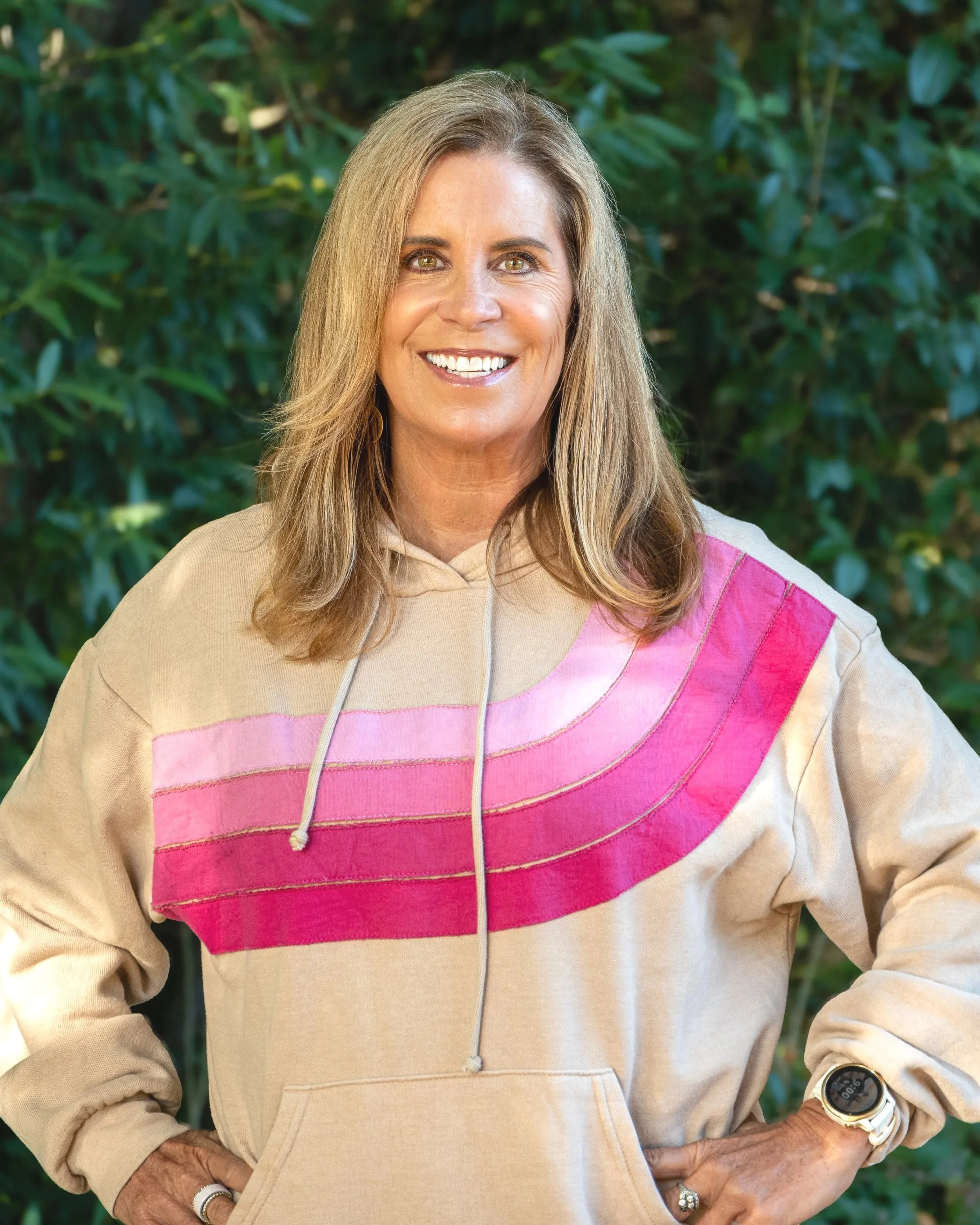A woman with long, wavy blonde hair and light skin smiling outdoors. She is wearing a beige hoodie with pink and purple stripes across the chest, a white wristwatch, and a ring on her right hand. She is standing in front of green foliage.