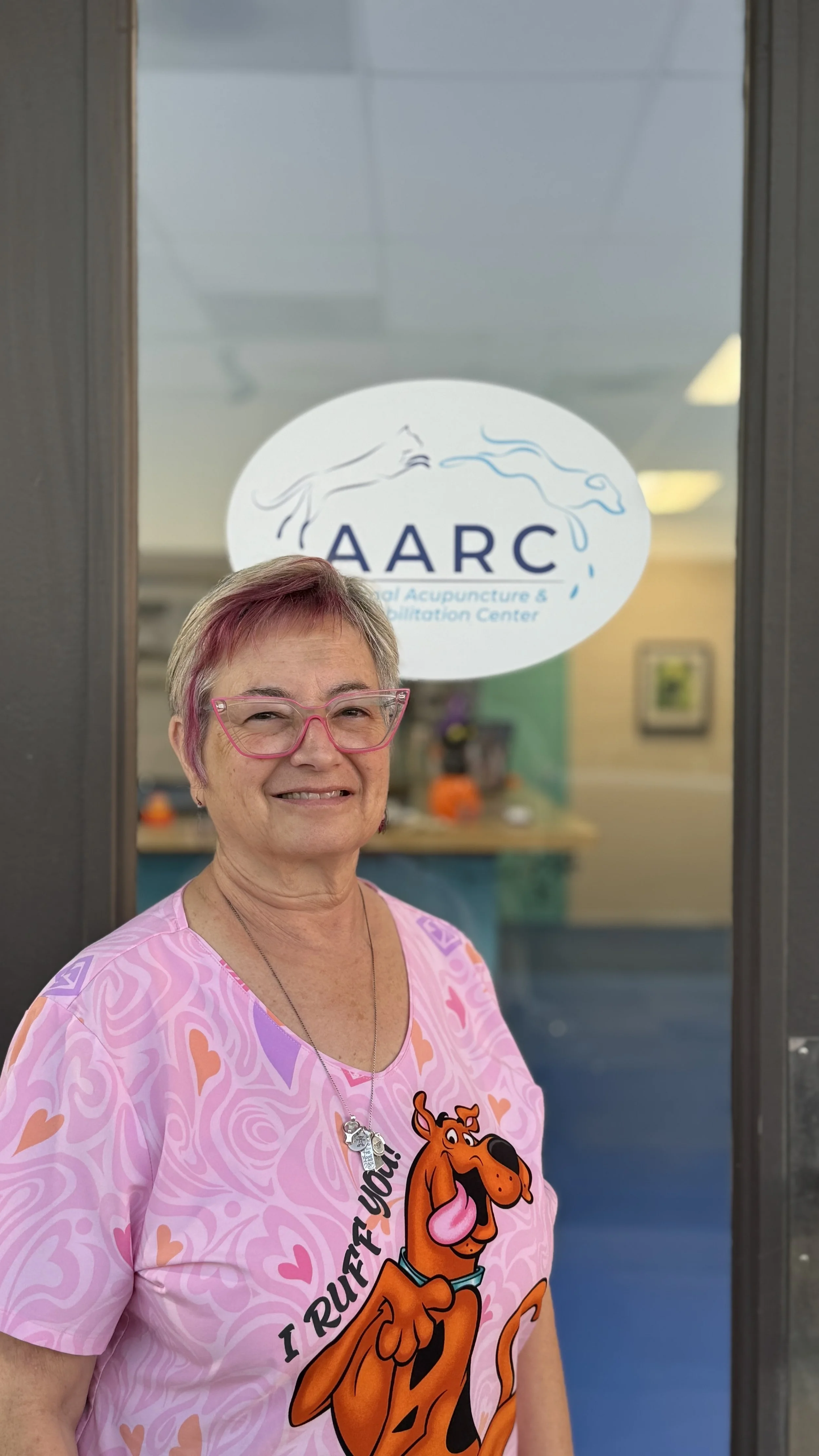A smiling woman wearing pink glasses and a pink T-shirt with Scooby-Doo on it standing in front of a window with a sign that reads 'AARC' for a holistic acupuncture and rehabilitation center.