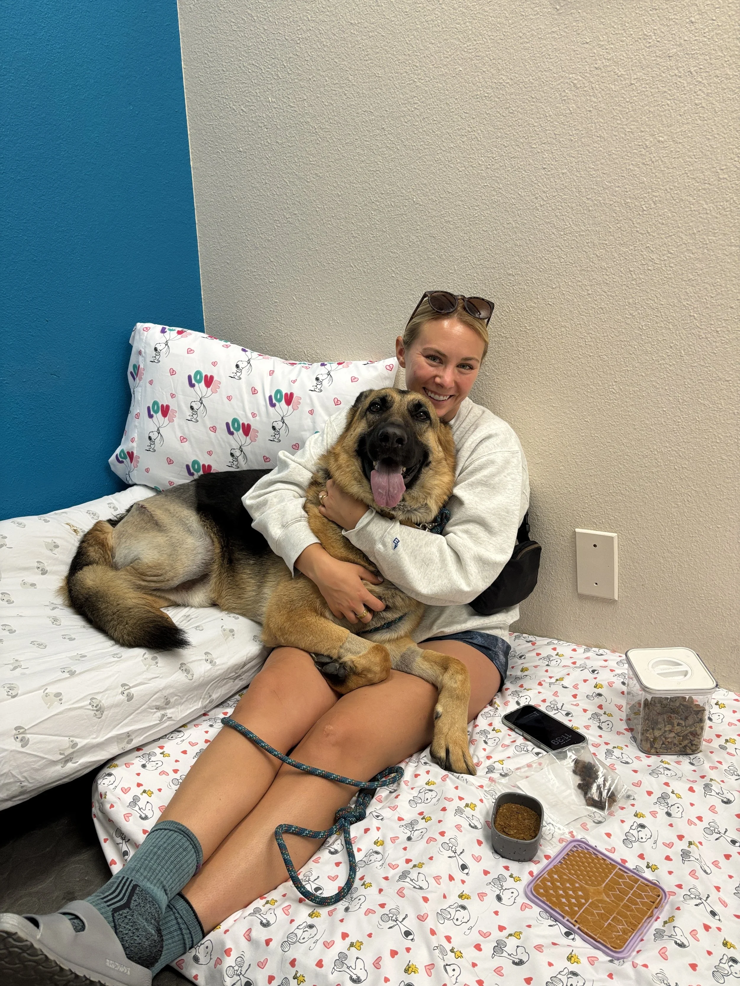 A woman sitting on a hospital bed hugging a large German Shepherd dog with a happy expression. The bed has Snoopy print sheets and a pillow with 'LOVE' and Snoopy illustrations. The woman is smiling, wearing sunglasses on her head and casual clothing