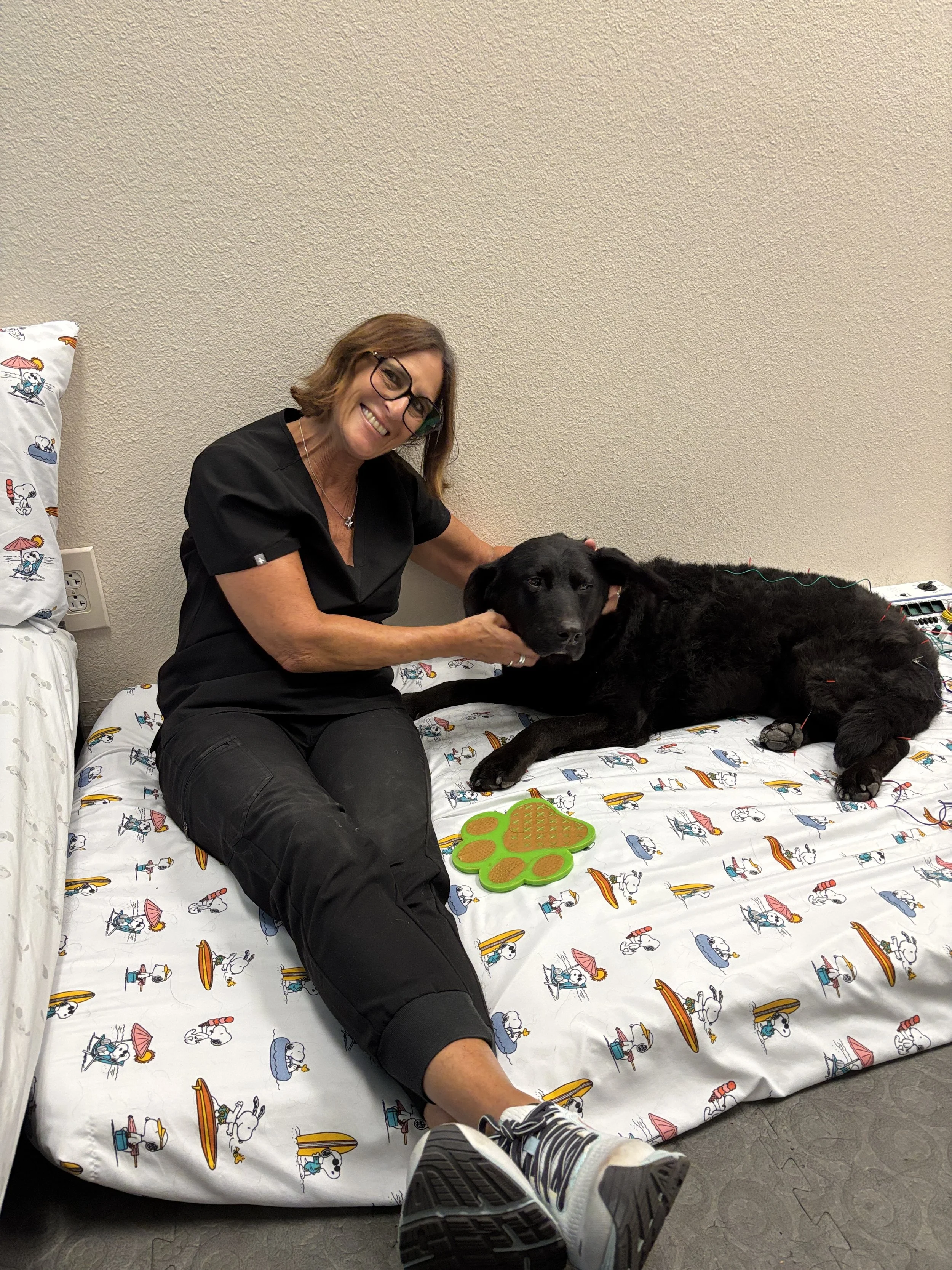A woman in medical scrubs sitting on a bed with Snoopy themed bedding, petting a black dog lying down beside her. The woman is smiling and wearing glasses, and the room has a neutral beige wall with medical equipment nearby.