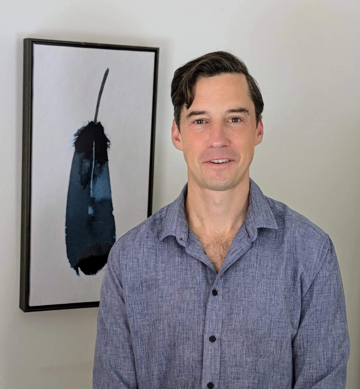 A smiling man with dark brown hair wearing a gray button-up shirt standing in front of abstract art on a white wall.