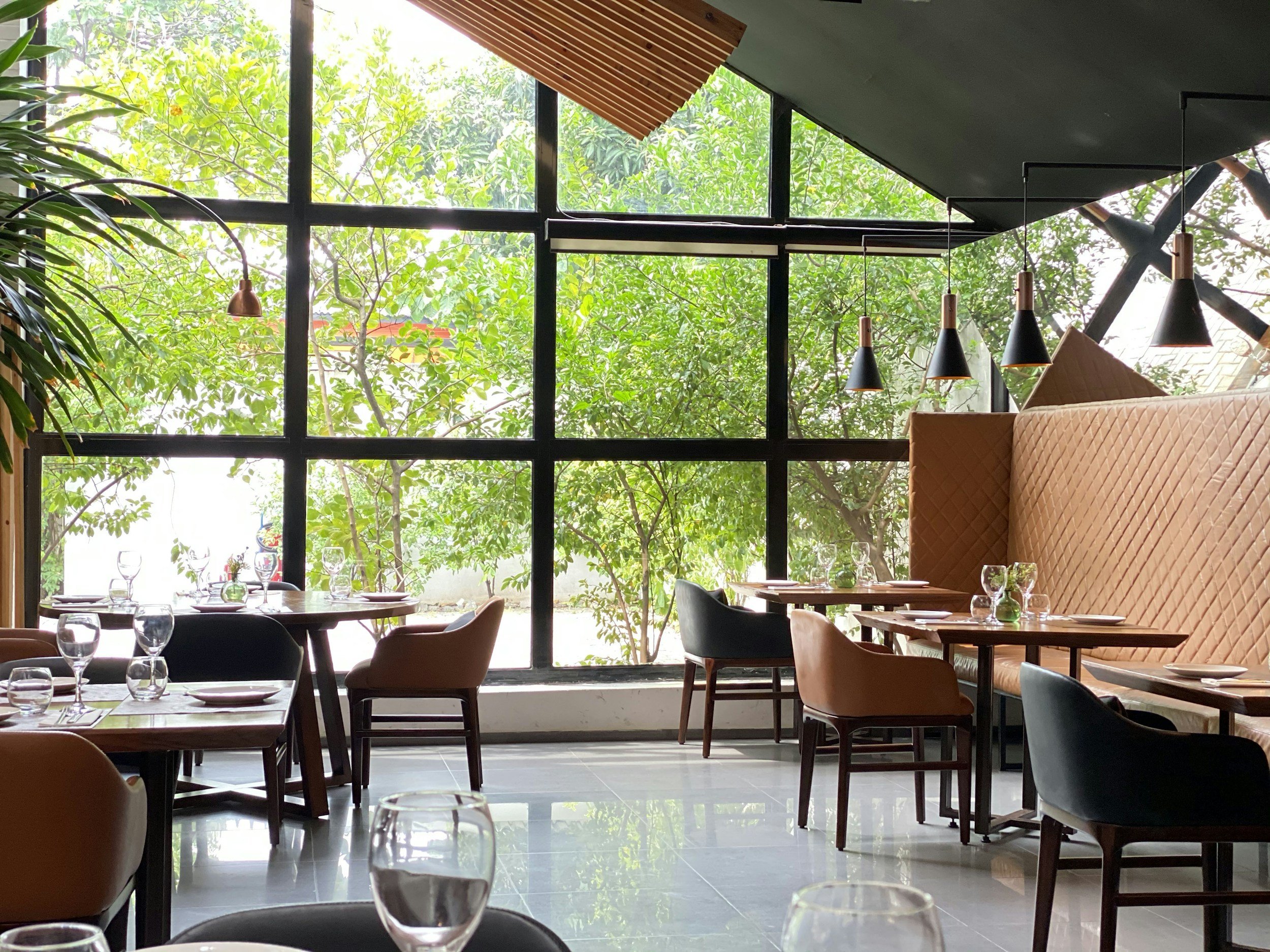 A restaurant dining area with tables set with wine glasses, plates, and utensils, large windows with green trees outside, modern black hanging lights, and cushioned upholstered seating.