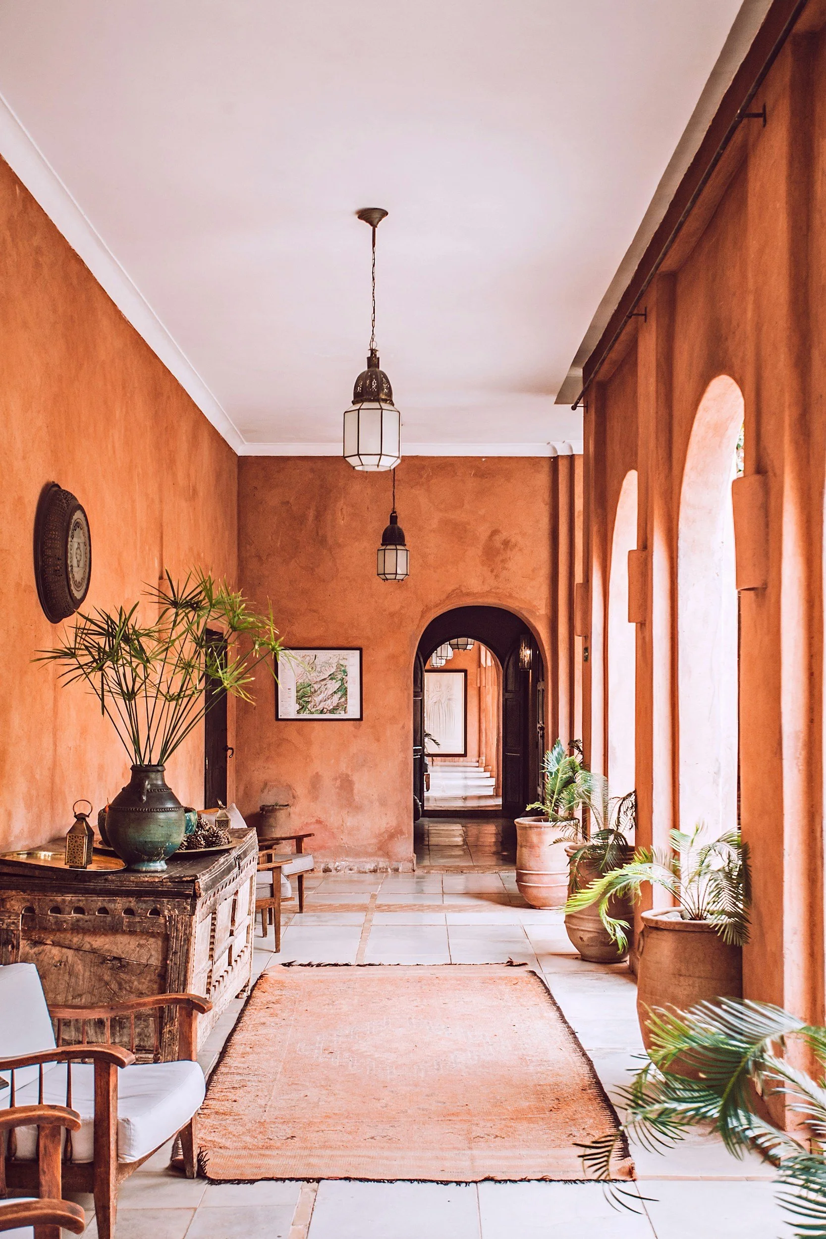 A hallway with warm orange and terracotta walls, wooden furniture, potted plants, and hanging light fixtures.