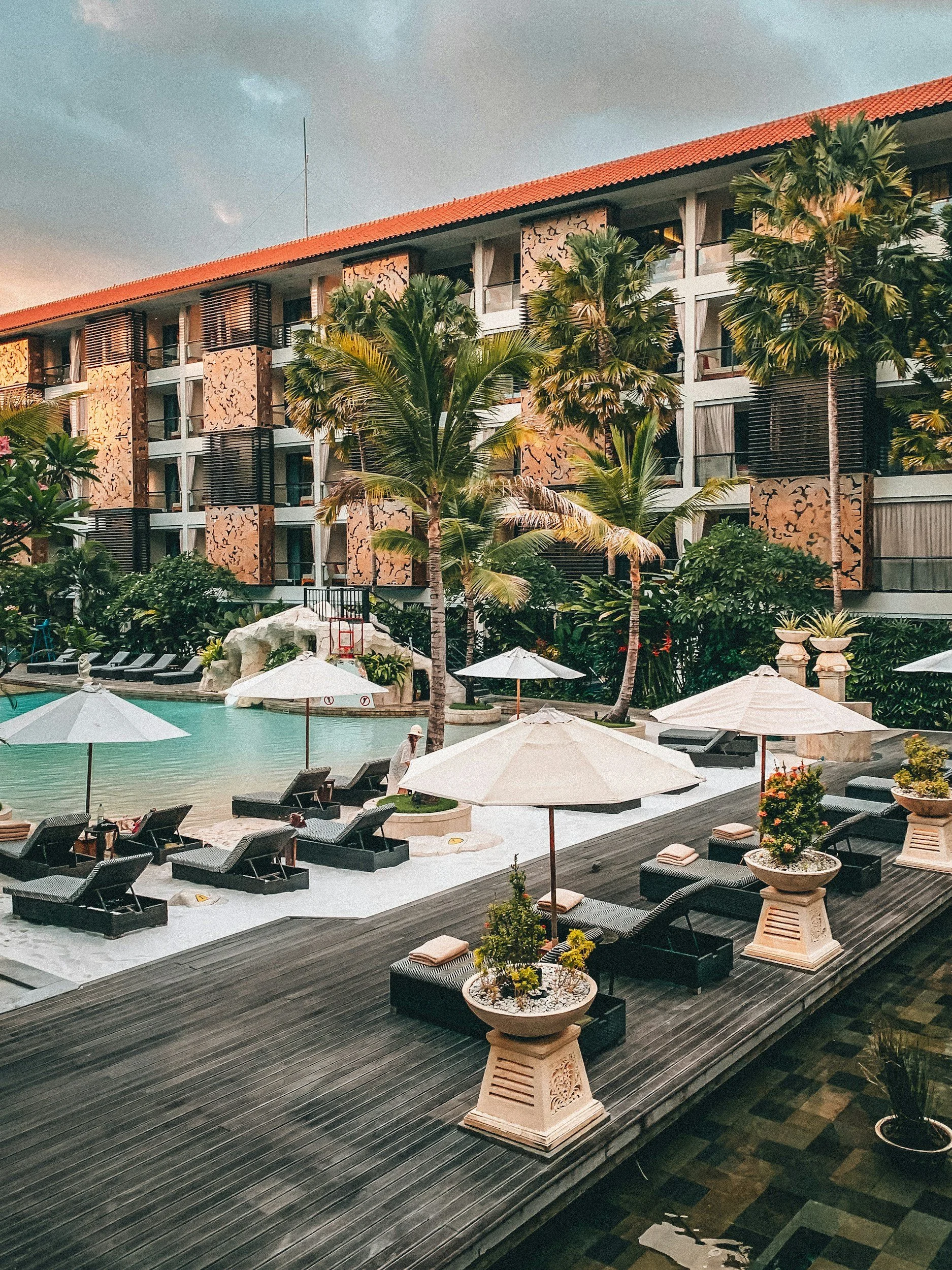 A view of a hotel pool area with lounge chairs, umbrellas, potted plants, and palm trees, with a hotel building in the background and a cloudy sky.