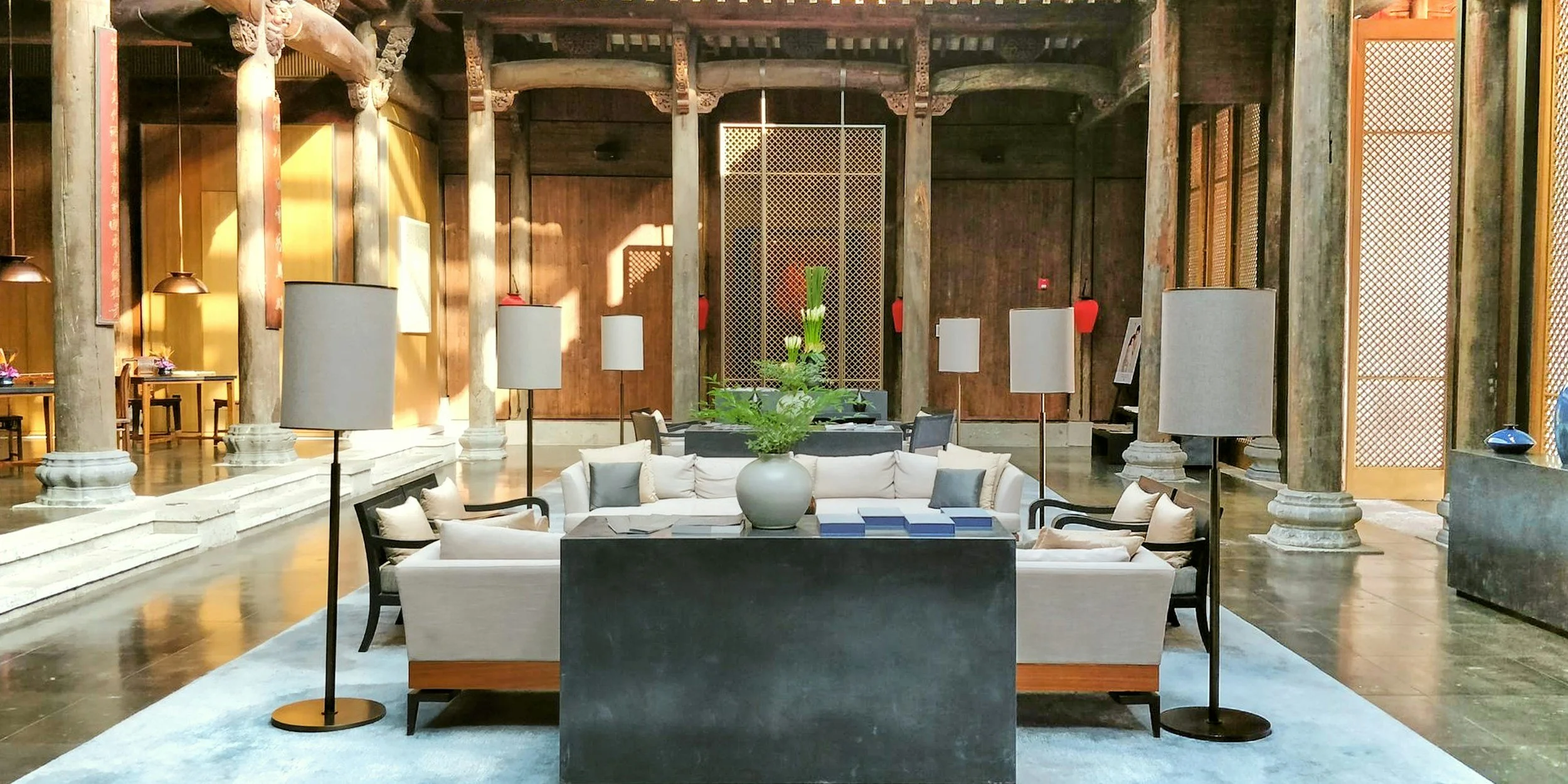 A spacious hotel lobby with a mix of modern and traditional Asian decor. It features a circular white sofa with gray cushions, black chairs, a large white vase with green plants, and tall floor lamps. Wooden columns and decorative panels with latticework are visible in the background, along with a reflective polished floor.