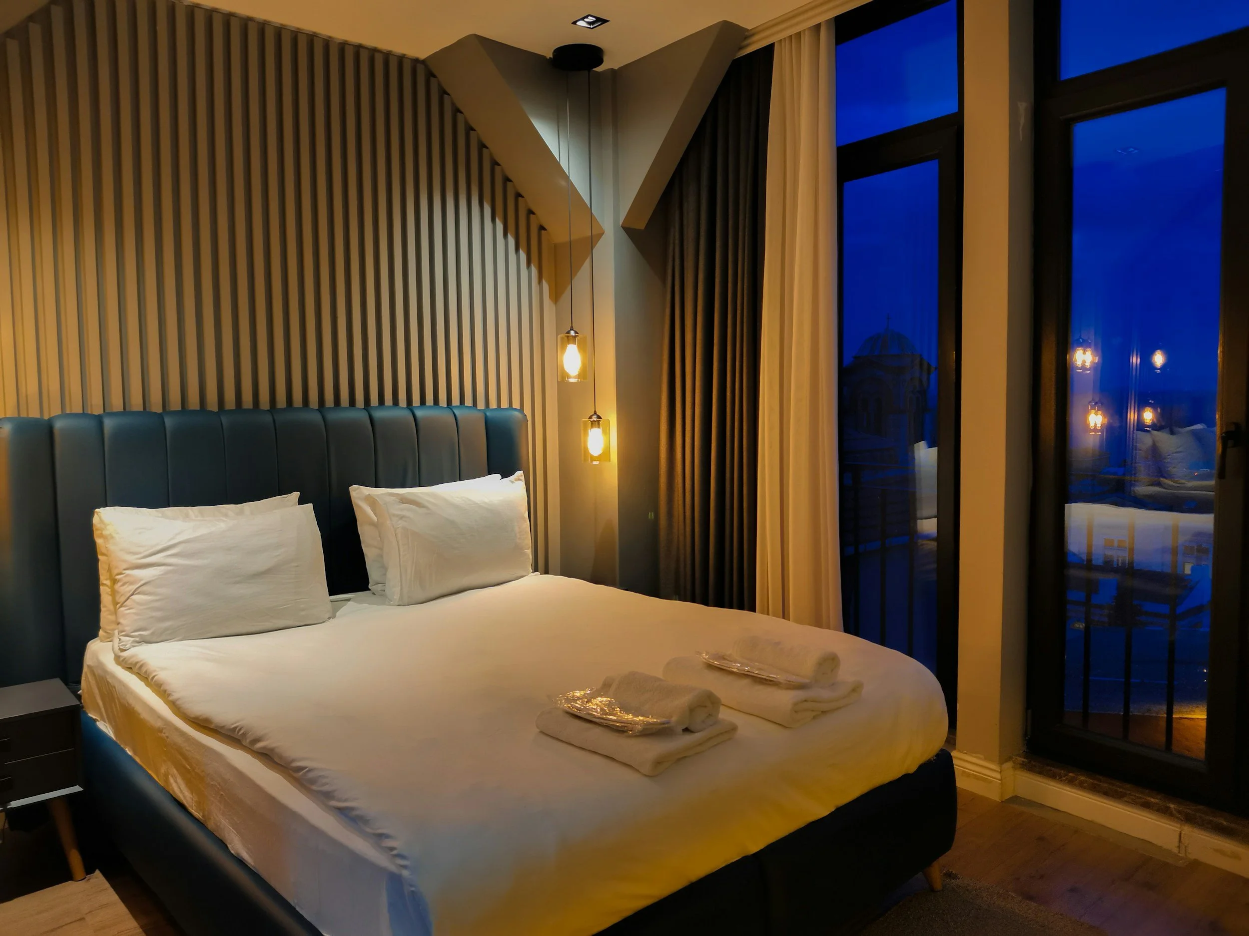 A neatly made bed with white sheets and pillows in a modern hotel room, lit by warm pendant lights, with large windows showing a cityscape at night.