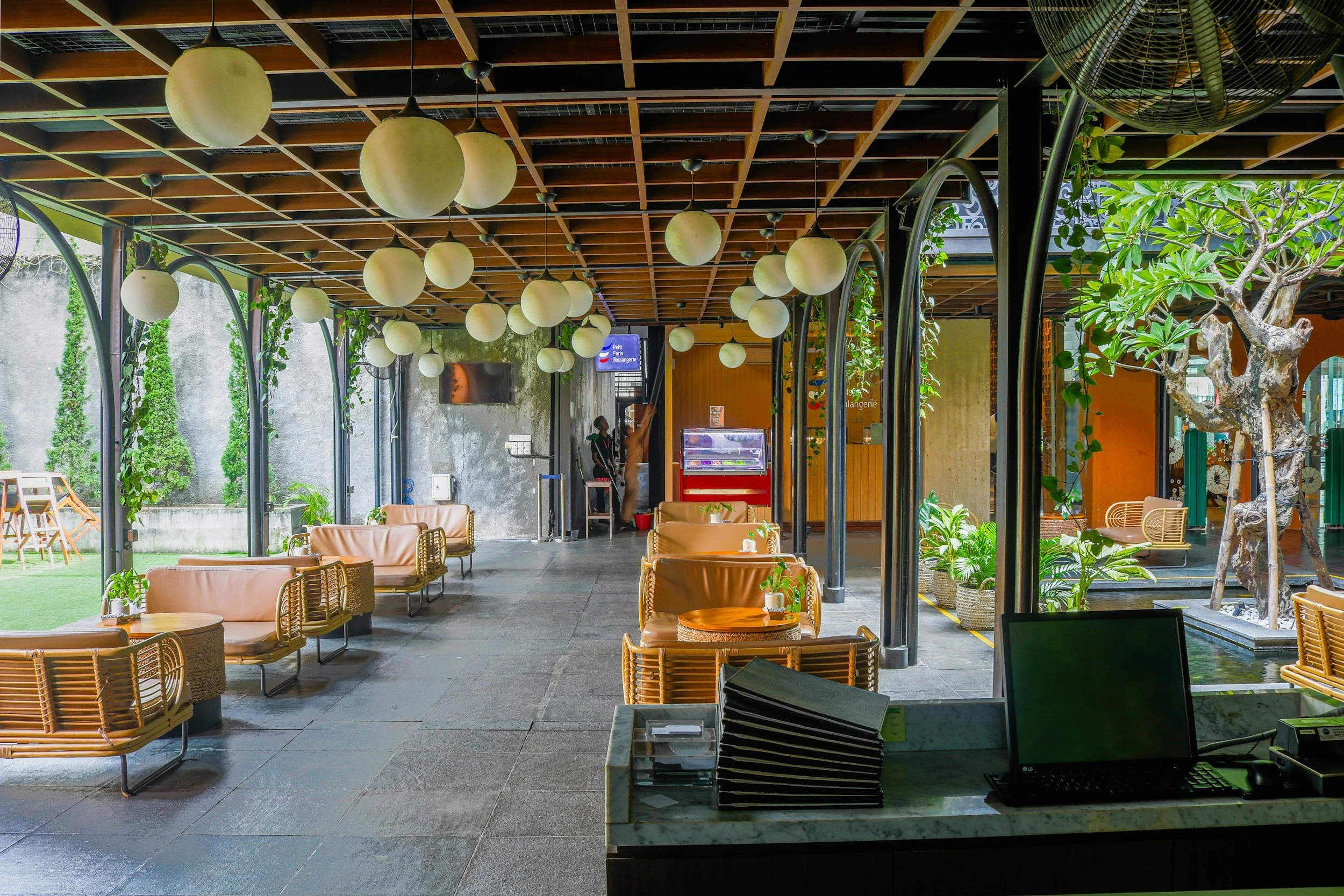 Interior of a modern cafe with hanging spherical lights, wooden ceiling grid, green plants, and outdoor view with tables, chairs, and a person in the background.
