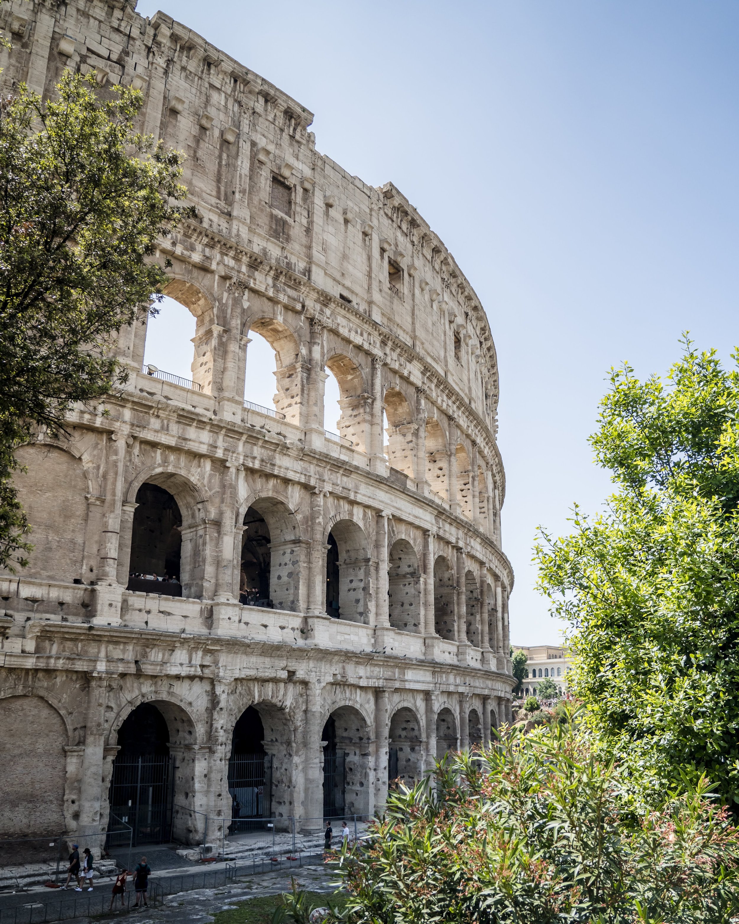 The Colosseum in Rome, Italy, with visitors walking outside and green trees in the foreground on a clear day.