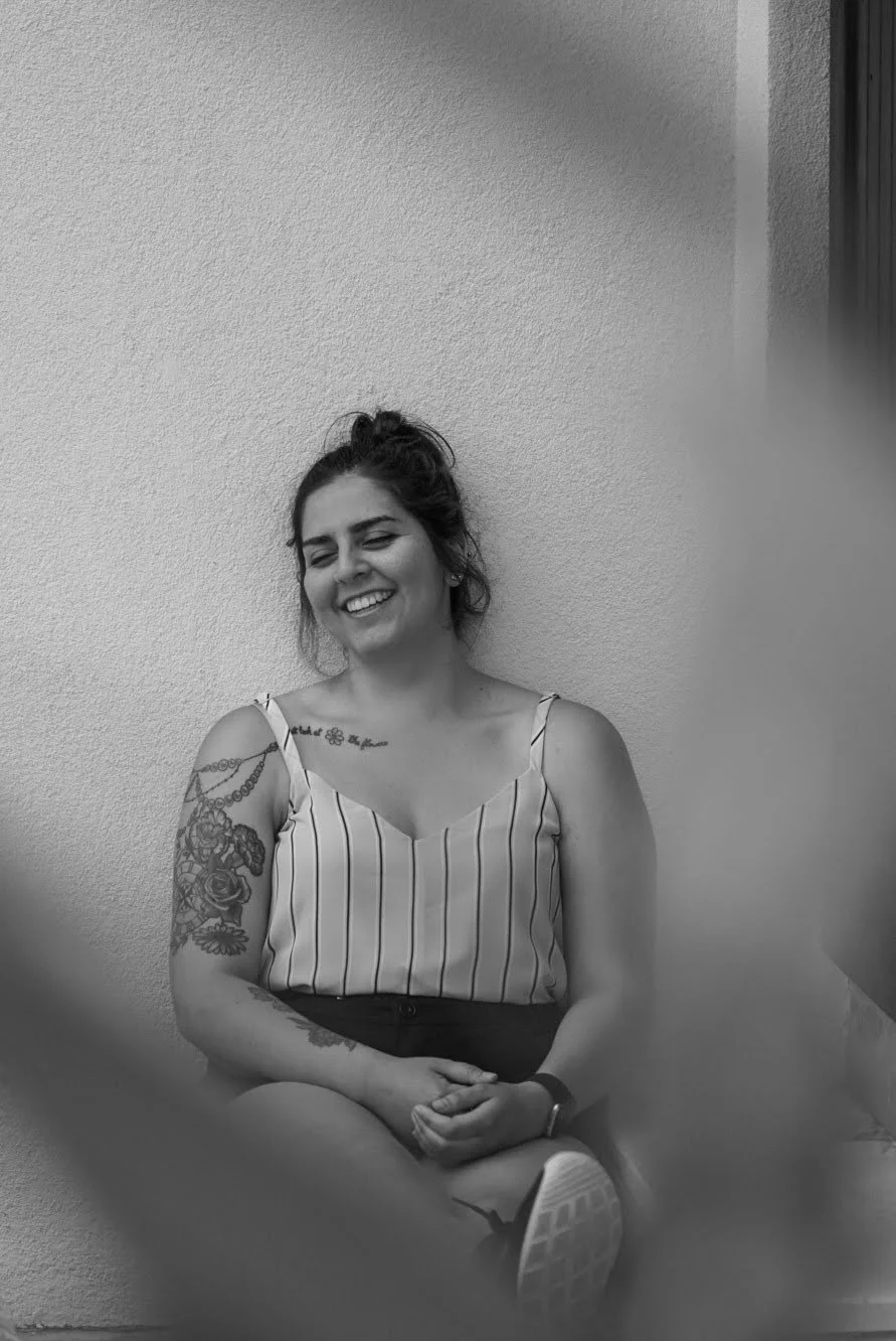 A woman with dark hair tied in a bun, wearing a striped sleeveless top, sitting against a plain wall, smiling with eyes closed, with visible tattoos on her left arm and a flower tattoo on her collarbone.