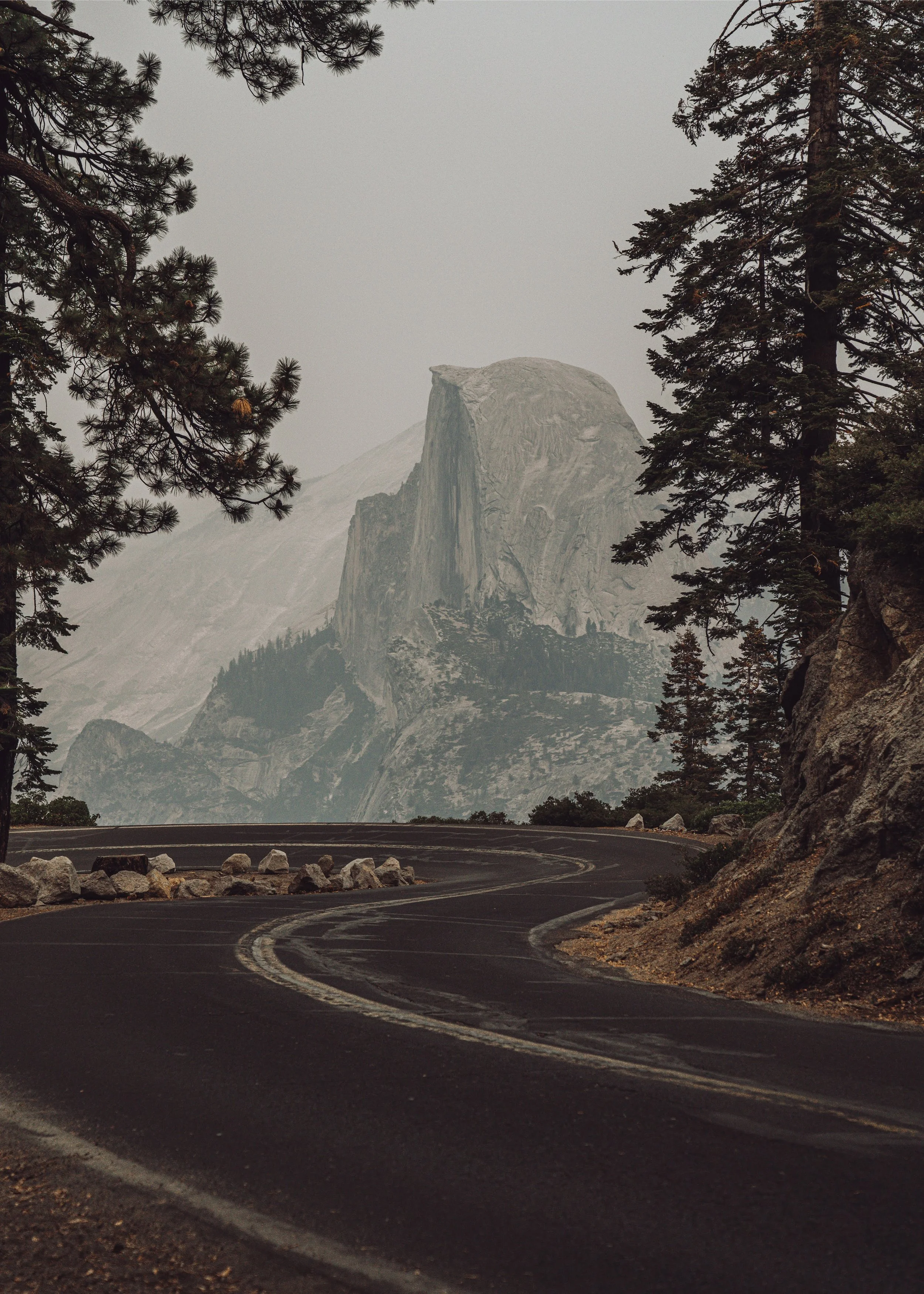 Curving mountain road with trees on both sides and a large granite mountain in the background under a cloudy sky.