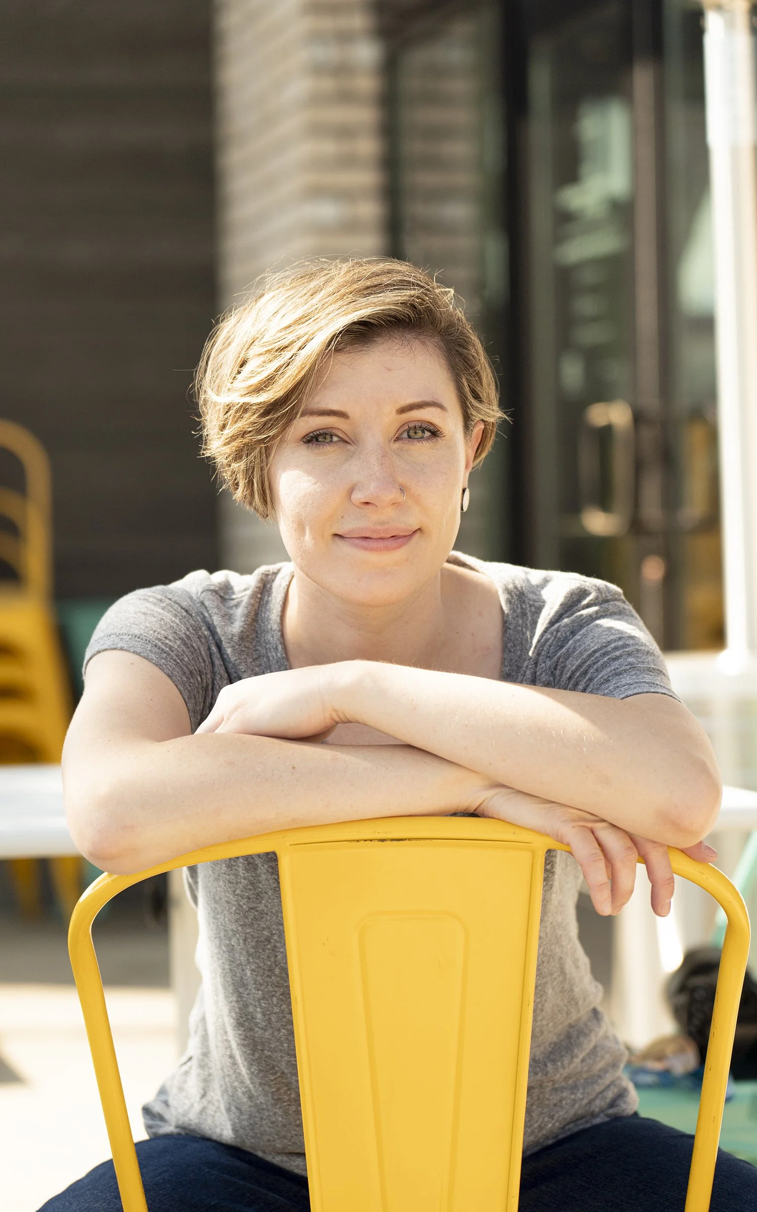 A young woman with short blonde hair and a nose ring, sitting outdoors behind a yellow chair, smiling at the camera.