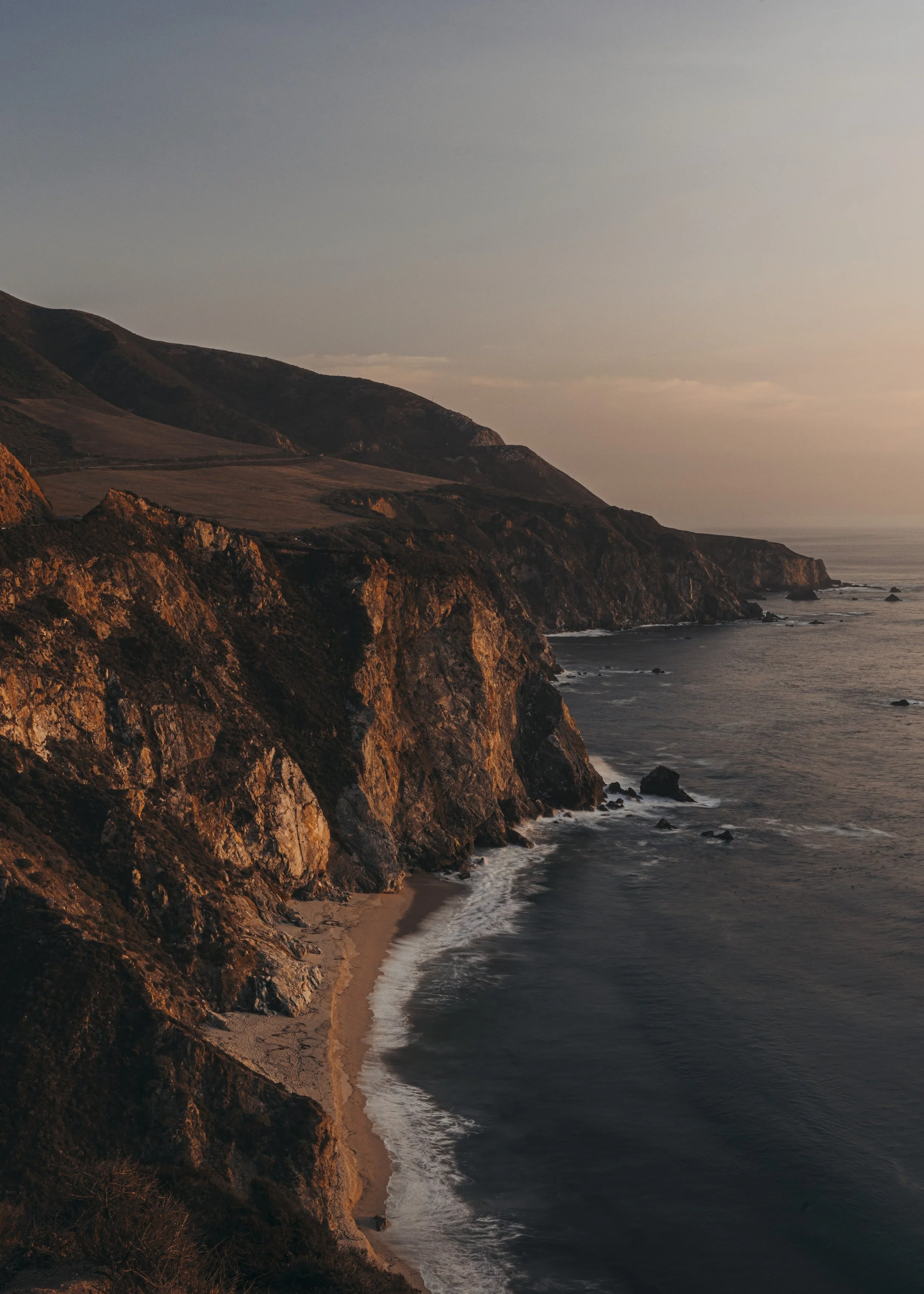 A rugged coastline at sunset with cliffs and a small beach at the base of the cliffs. The ocean waves gently hit the shore, and the sky is tinged with warm colors from the setting sun.