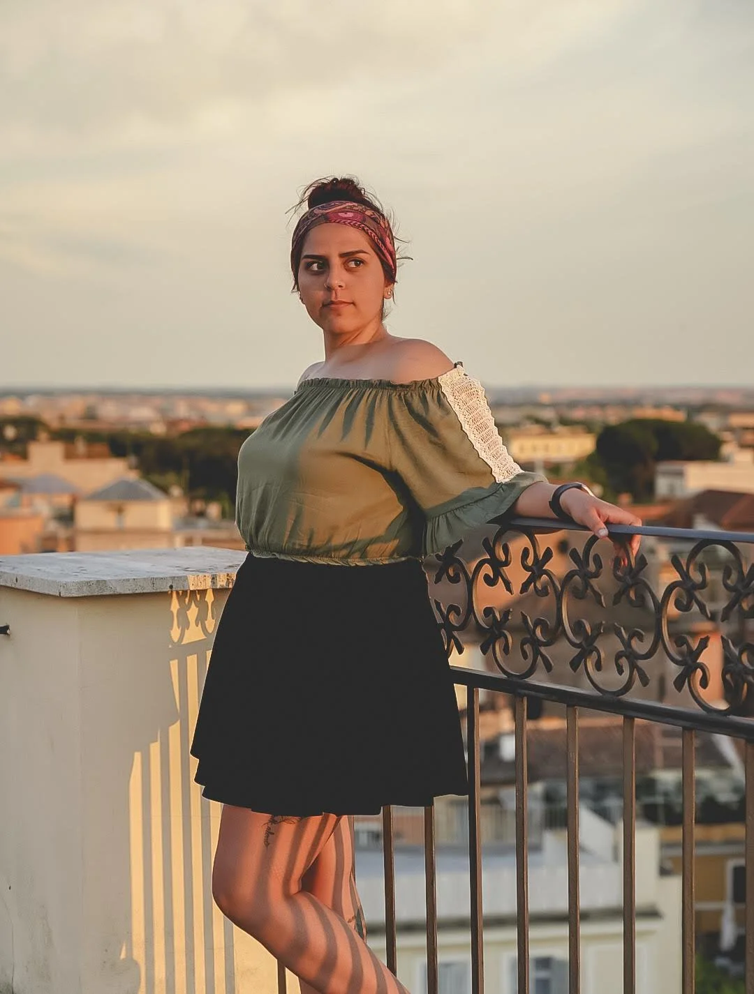 A woman wearing an off-shoulder blouse and a black skirt standing on a rooftop balcony during sunset, with a cityscape in the background.