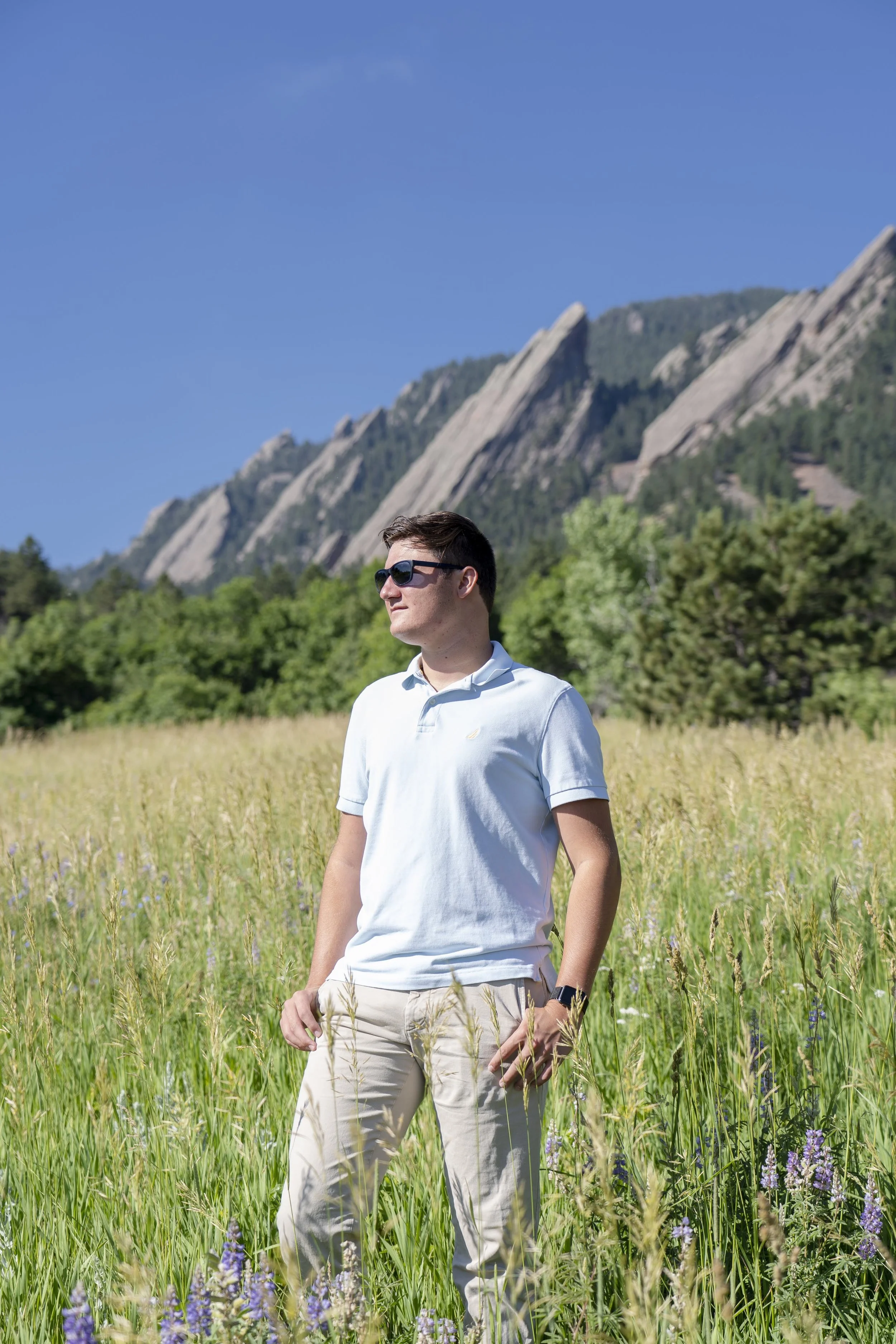 A man wearing sunglasses, a white polo shirt, and beige pants is standing in a field of tall grass and wildflowers with mountains in the background under a blue sky.