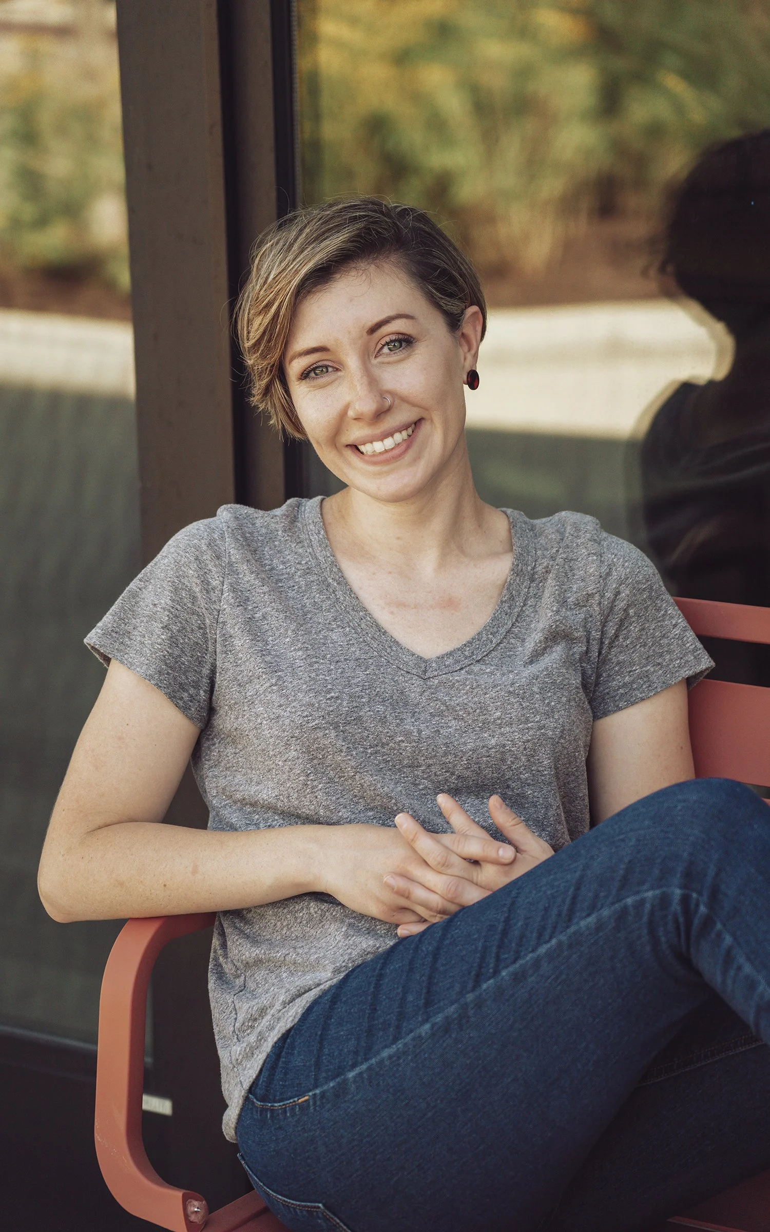 A woman with short hair, wearing a gray t-shirt and jeans, sitting on a pink chair next to a window with a blurred outdoor background, smiling at the camera.