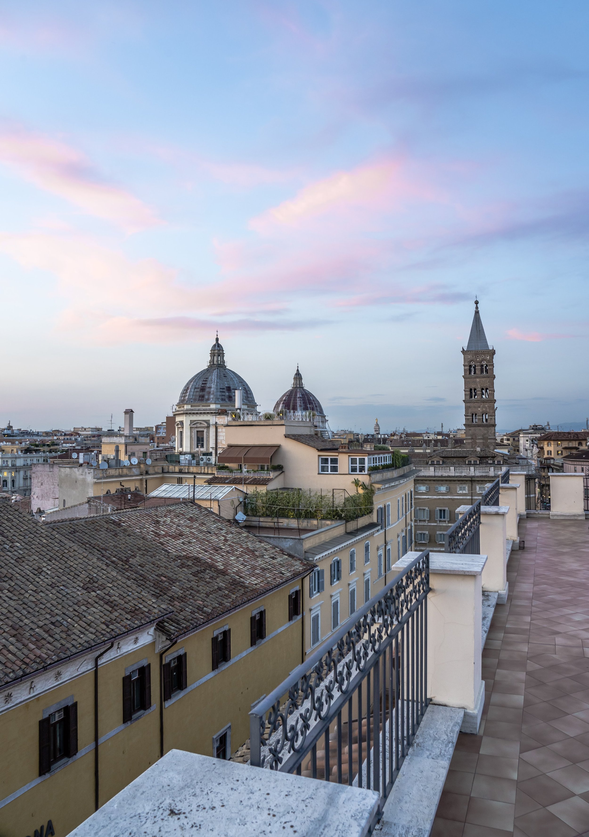 Cityscape of Rome, Italy with domed church buildings, tower, and rooftops under a pink and blue sky seen from a terrace with decorative railing.