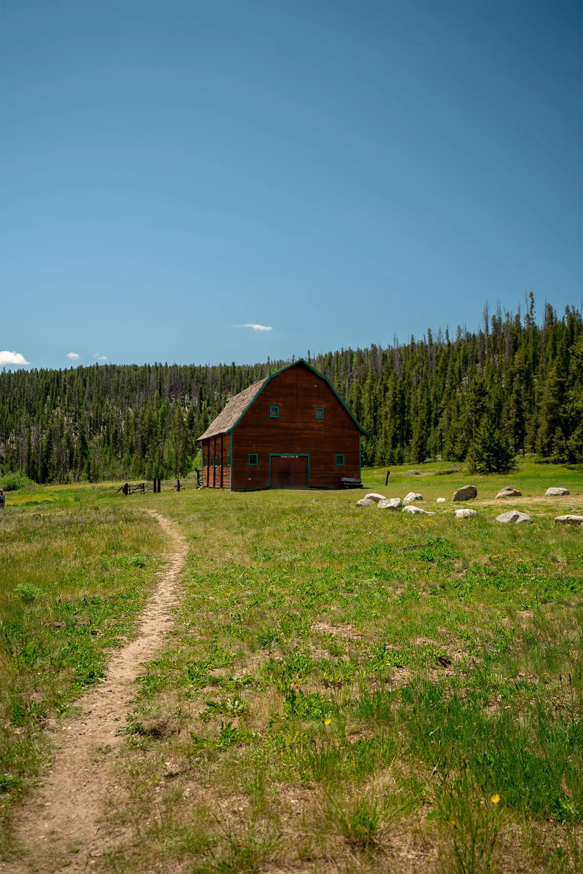 A dirt path leading to a brown barn with green trim, surrounded by grass, stones, and trees under a clear blue sky with a few clouds.