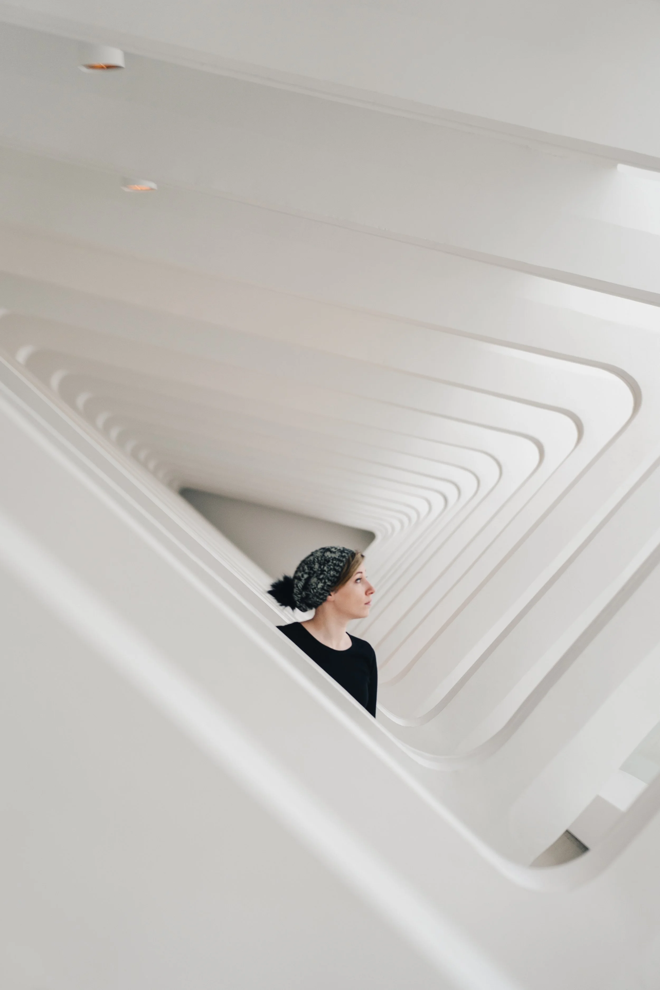 A woman with a black top and patterned headband looking to the right, standing inside a modern, spiral white staircase with curved railings.