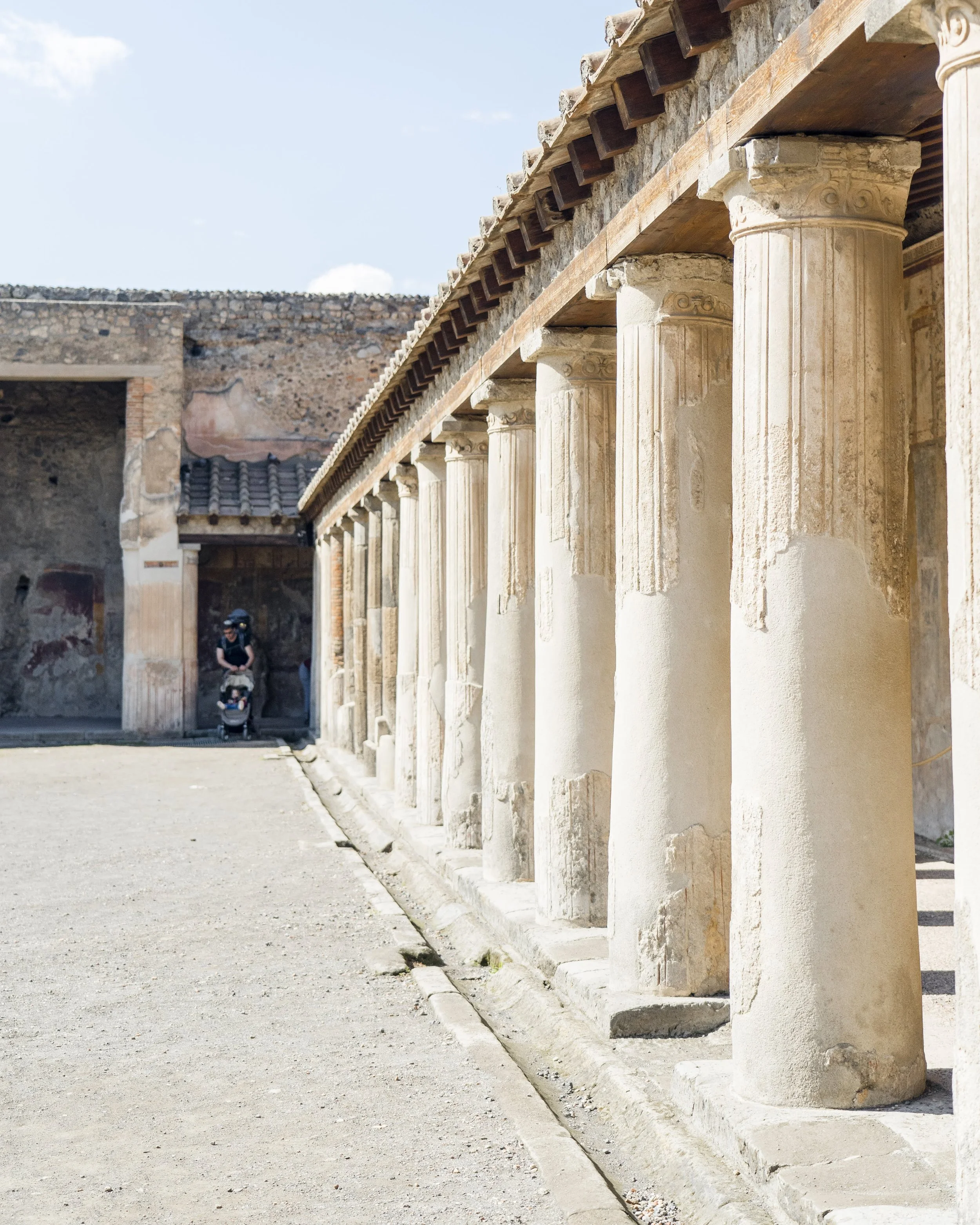 Ancient ruins with a row of classical columns made of stone or marble, supporting a wooden roof. The ruins are outdoor, with a clear sky and a few clouds visible. There is a person pushing a stroller in the background near the ruins.