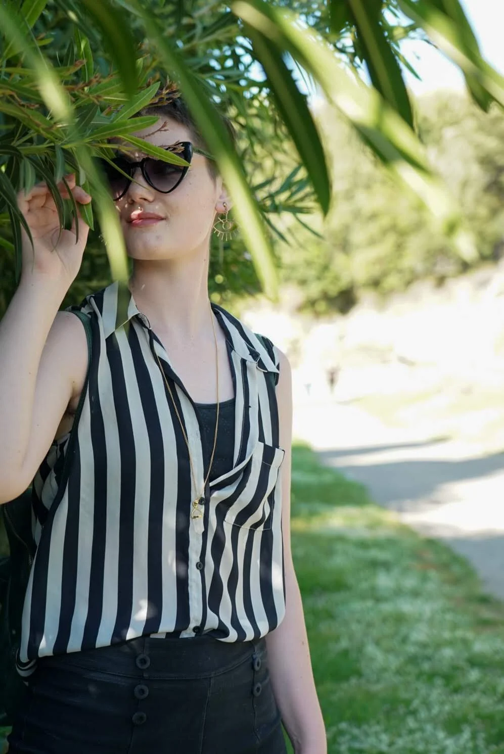 A woman with short hair, wearing black sunglasses, striped black and white sleeveless blouse, black pants, and gold earrings, standing outdoors under a tree with green leaves, smiling slightly while adjusting her glasses.