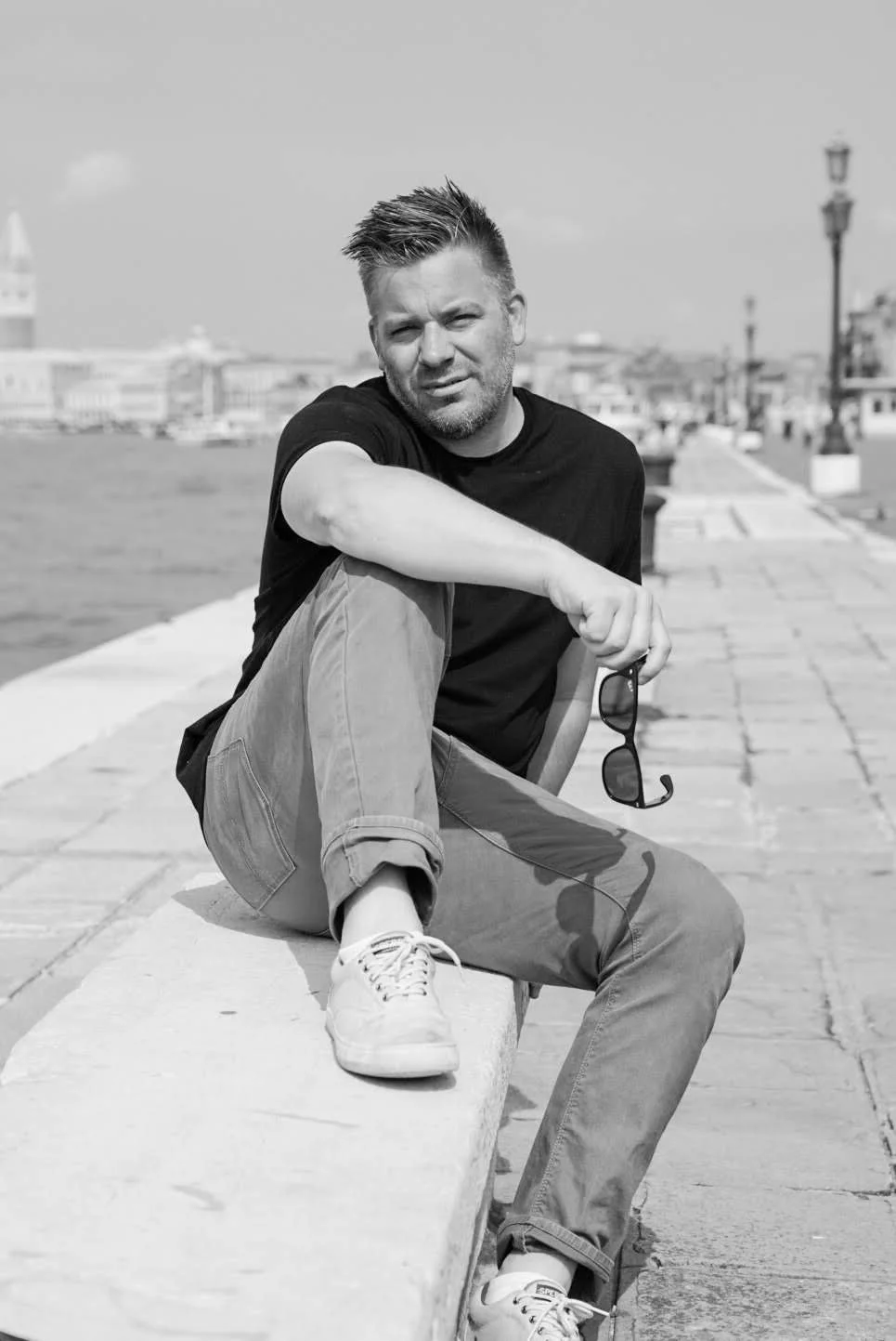 A man sitting on a ledge by the water, holding sunglasses, in an outdoor setting, with a cityscape in the background.